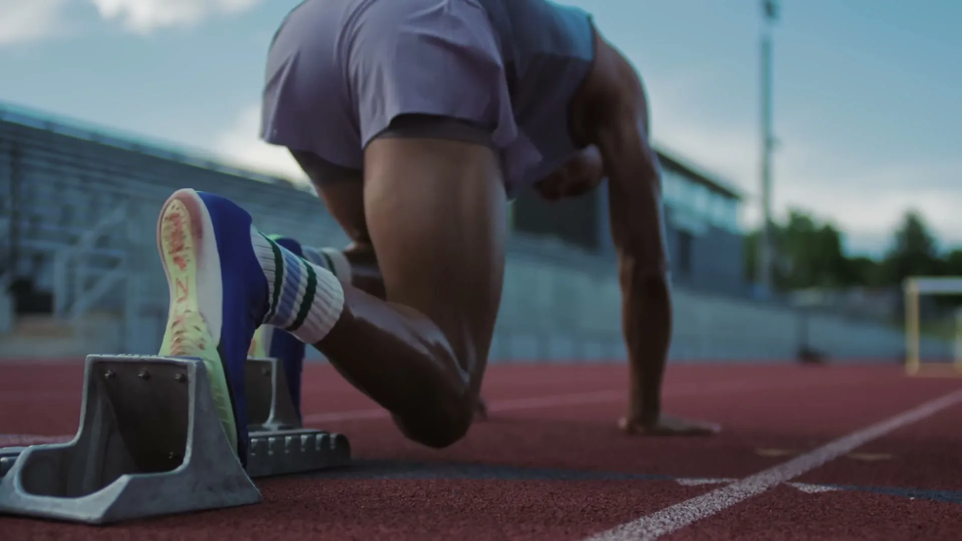 A male athlete in athletic wear is in a starting block on a red track, preparing to run at an outdoor stadium.