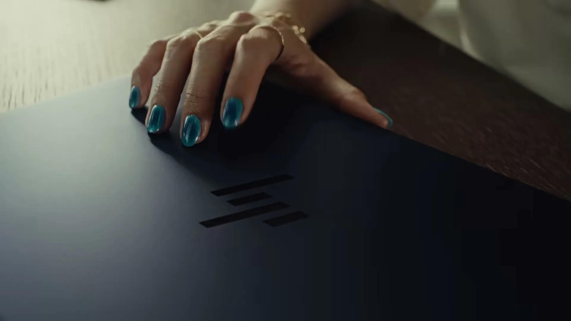 A woman's hand with teal nail polish rests on a dark laptop with the HP logo, on a wooden table.