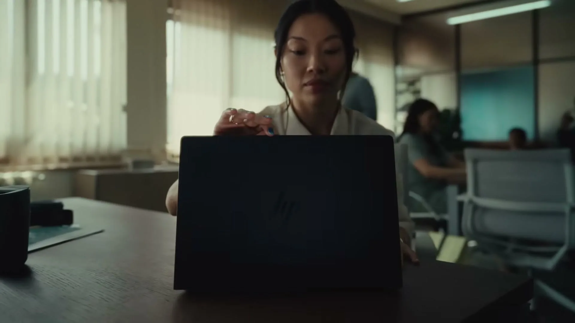 A young Asian woman in a white shirt opens a black HP laptop on a wooden desk in a busy office during the day.