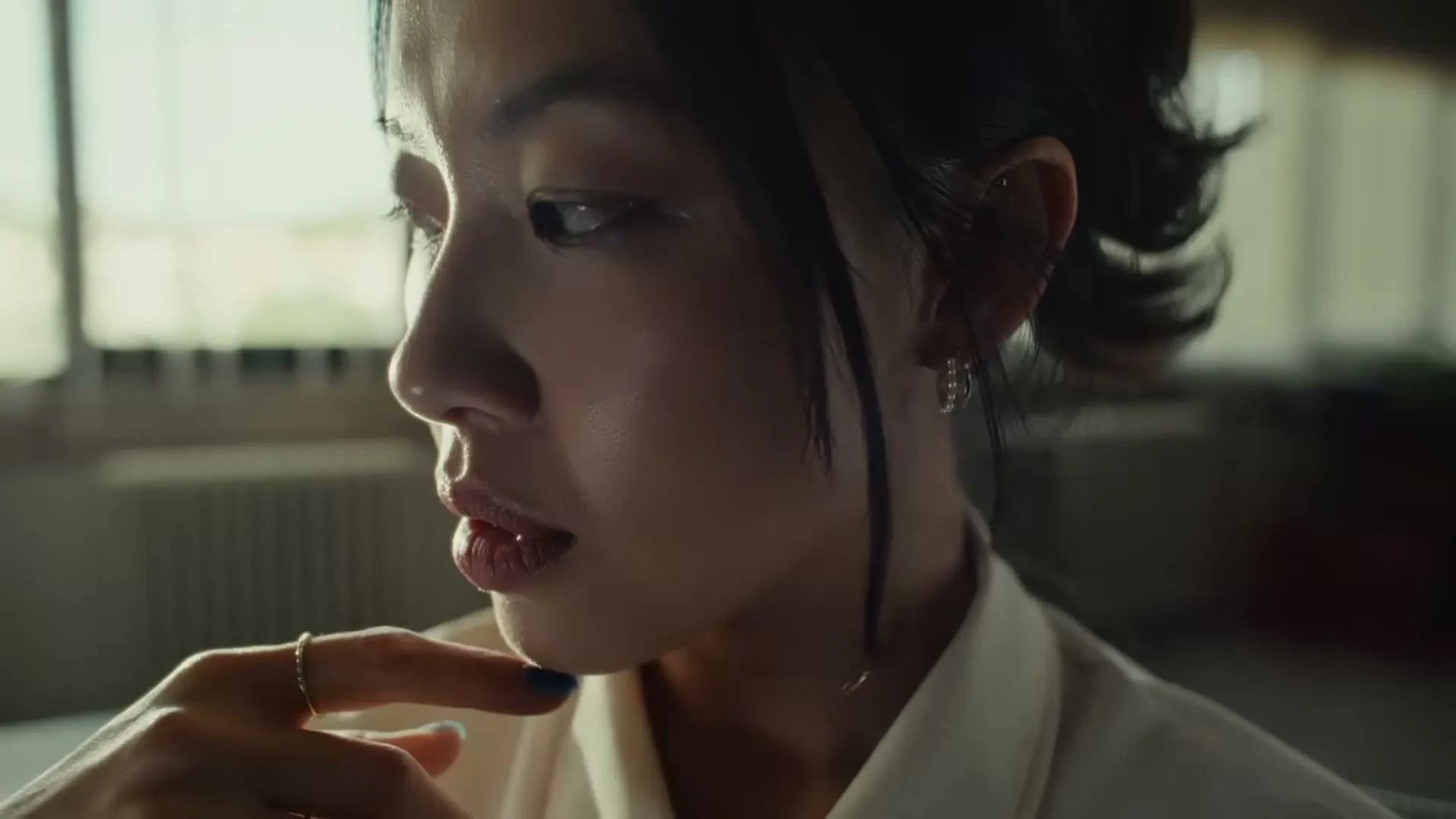 A young woman with dark hair and a white shirt looks down thoughtfully, touching her chin with a finger, in an interior setting.