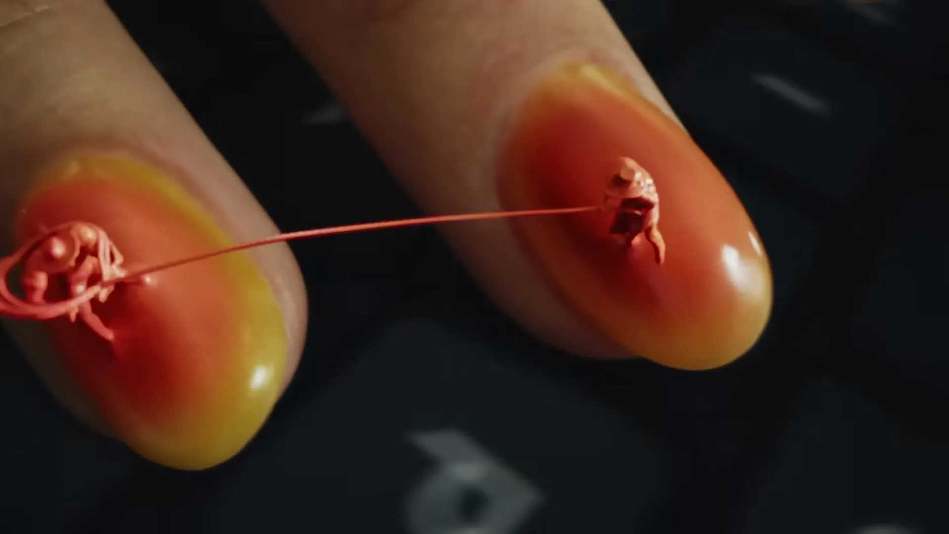 Two fingers with painted nails have tiny red figures on them, one figure pulls a string from the other, on a dark keyboard.