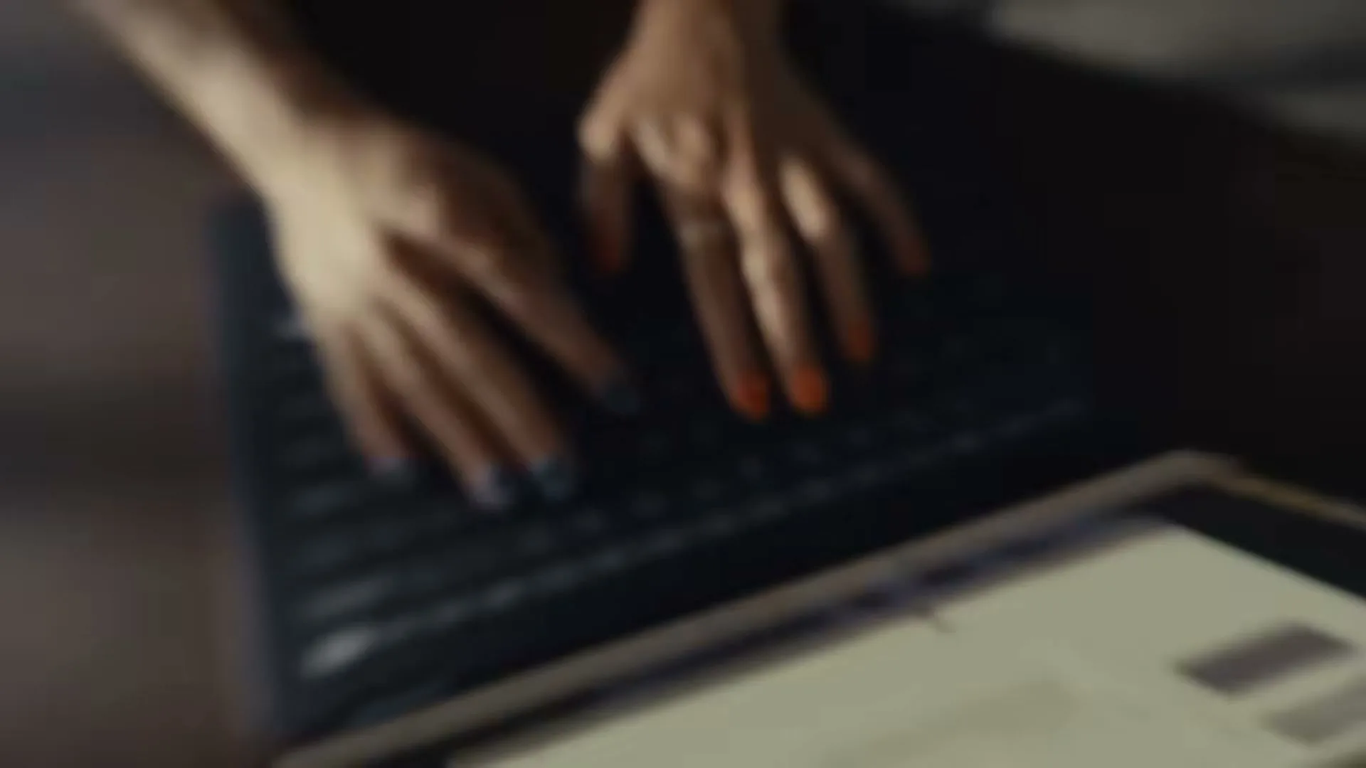 A woman's hands with colorful nail polish type on a laptop keyboard in a dimly lit room.