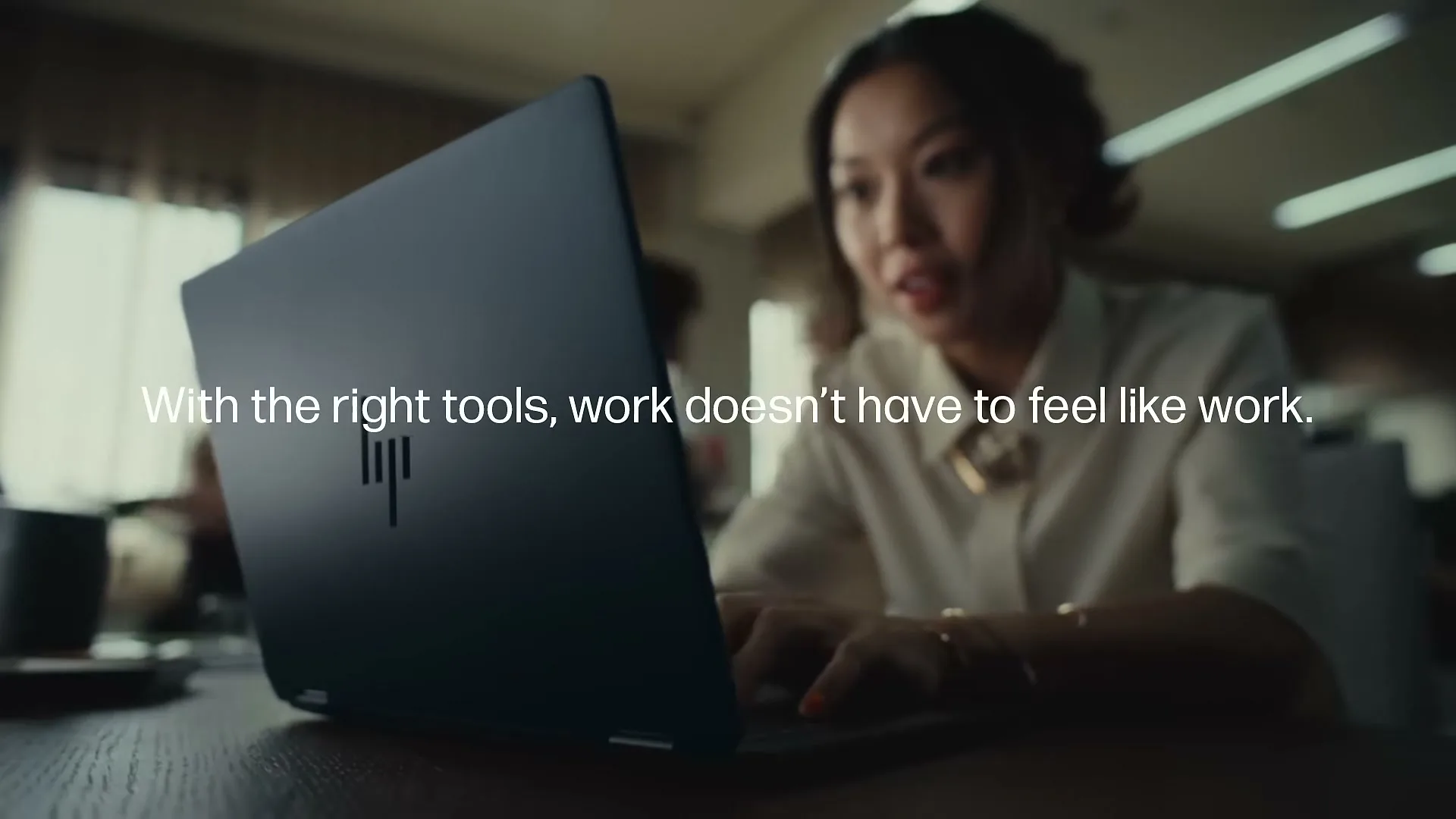A young Asian woman in a white shirt works on a laptop at a desk in a busy office, with text overlayed on the screen.