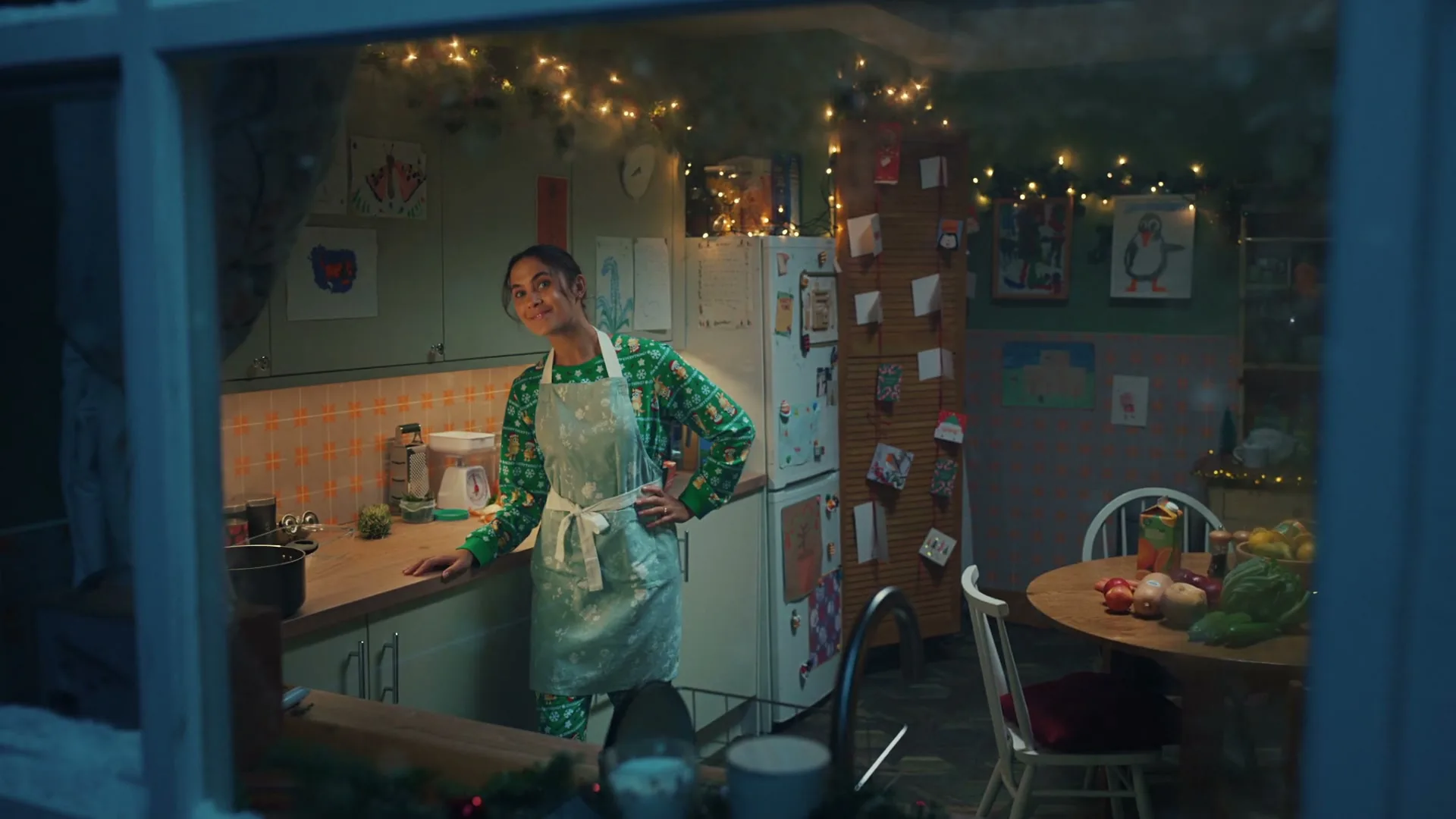 A woman in Christmas pajamas and an apron stands in a festive kitchen, looking out the window with a slight smile.