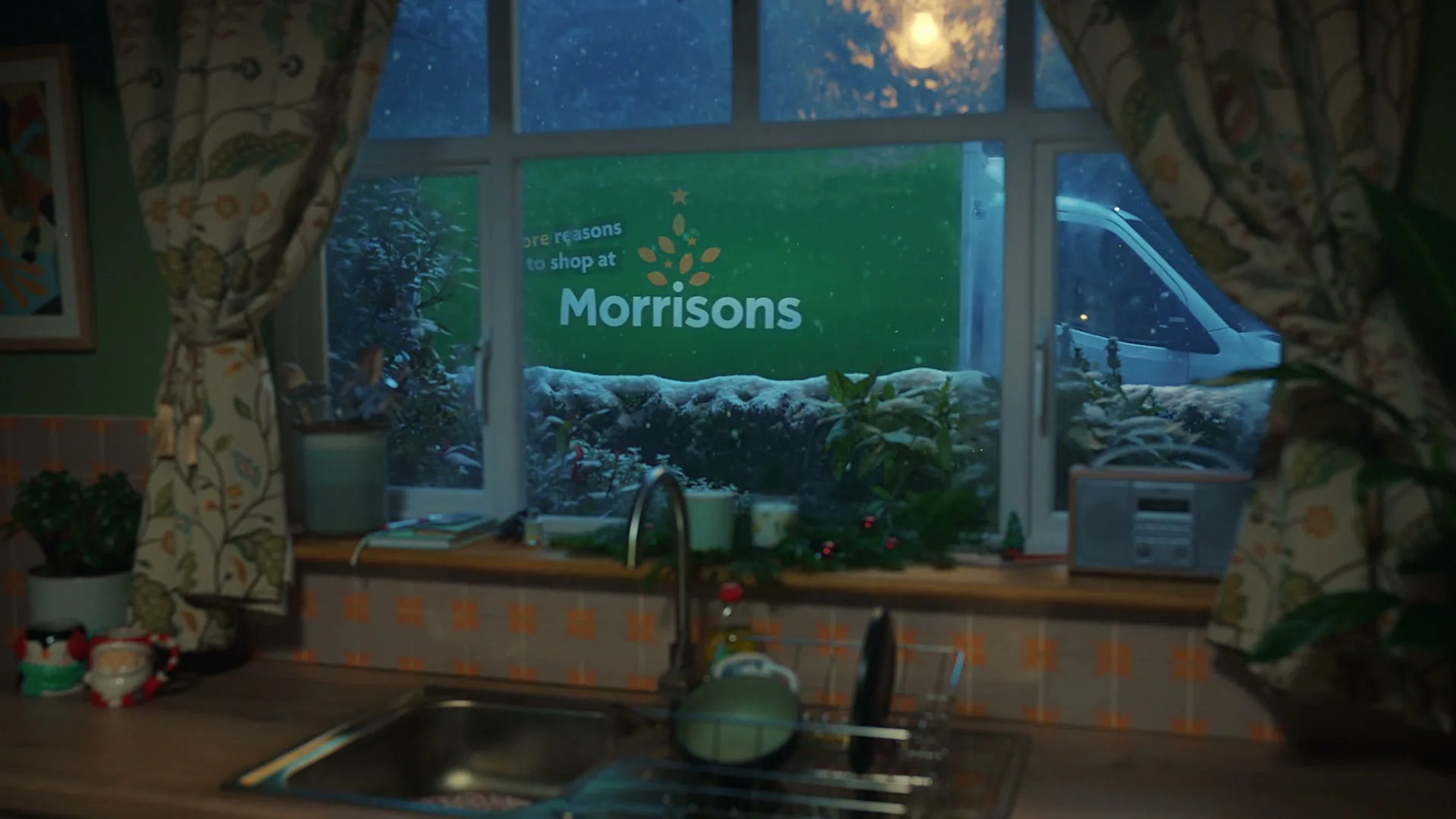 A Morrisons delivery truck is seen through a kitchen window on a snowy night, with the Morrisons logo visible on the side of the truck.