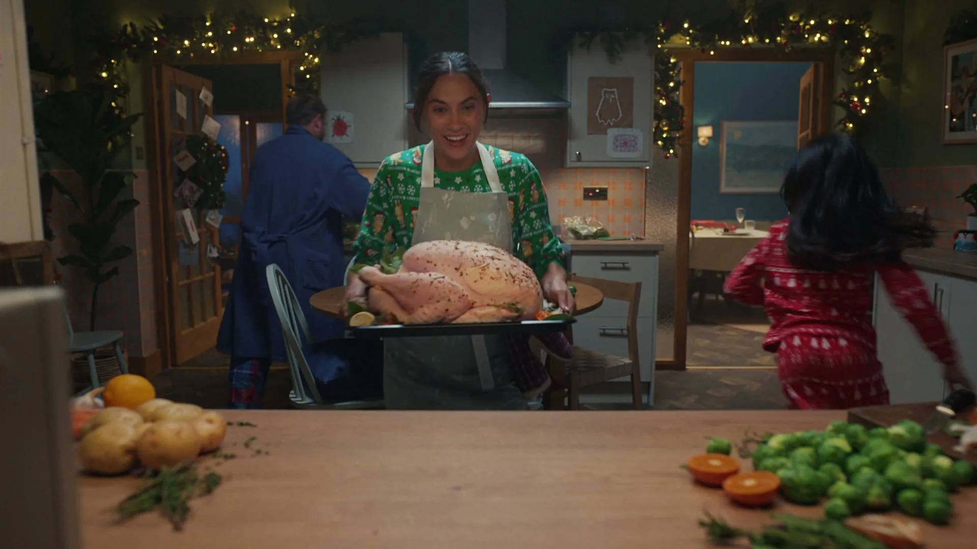 A woman in a Christmas sweater and apron carries a raw turkey on a tray, placing it on a kitchen counter while a child runs past and a man is in the background.