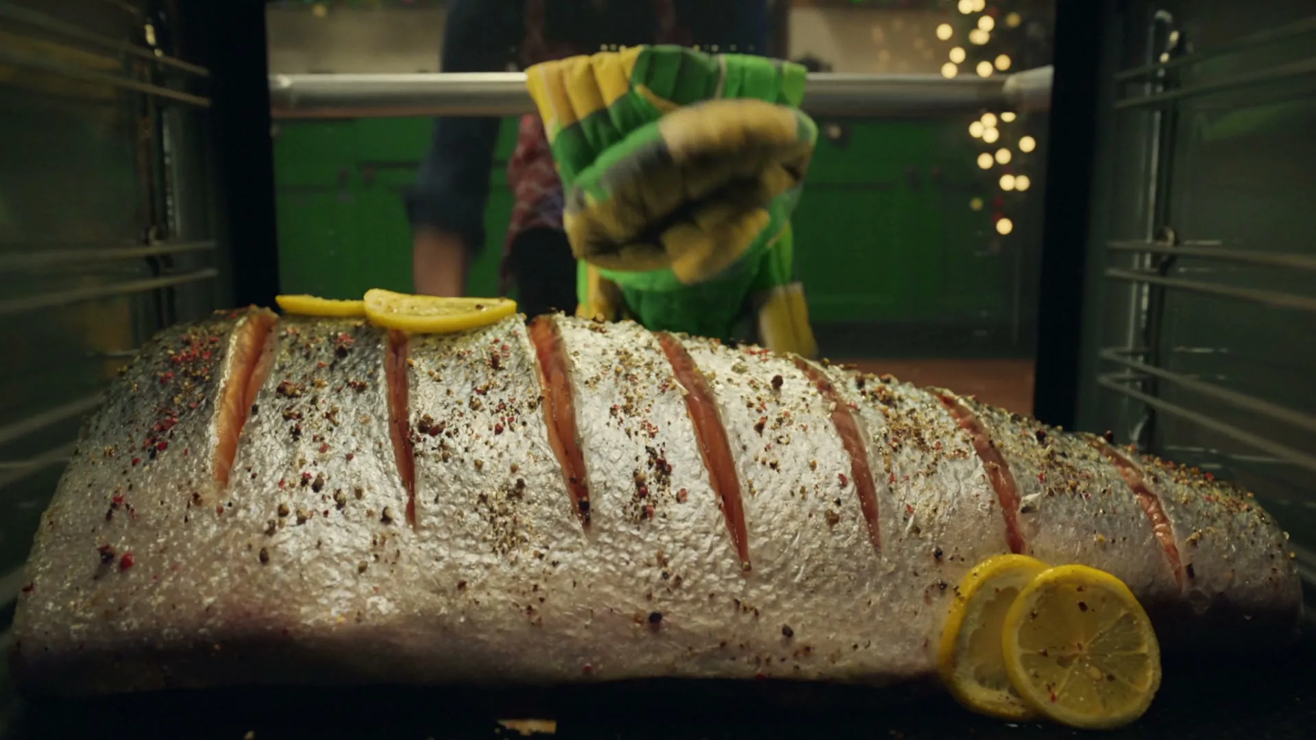 A man in an apron puts a large, seasoned salmon with lemon slices into an oven, preparing a meal in a festive kitchen.