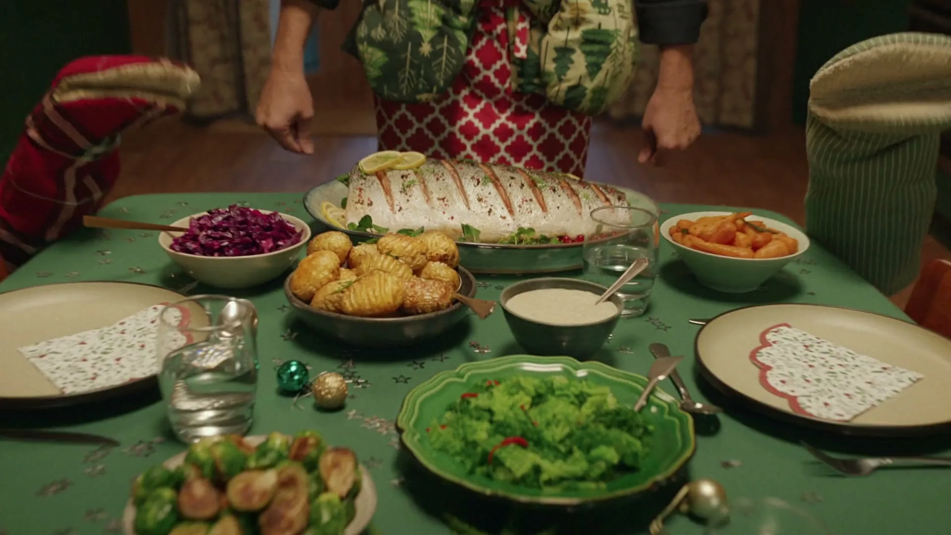 A man in a festive apron places a large cooked fish on a table laden with Christmas dinner, then spreads his arms wide.
