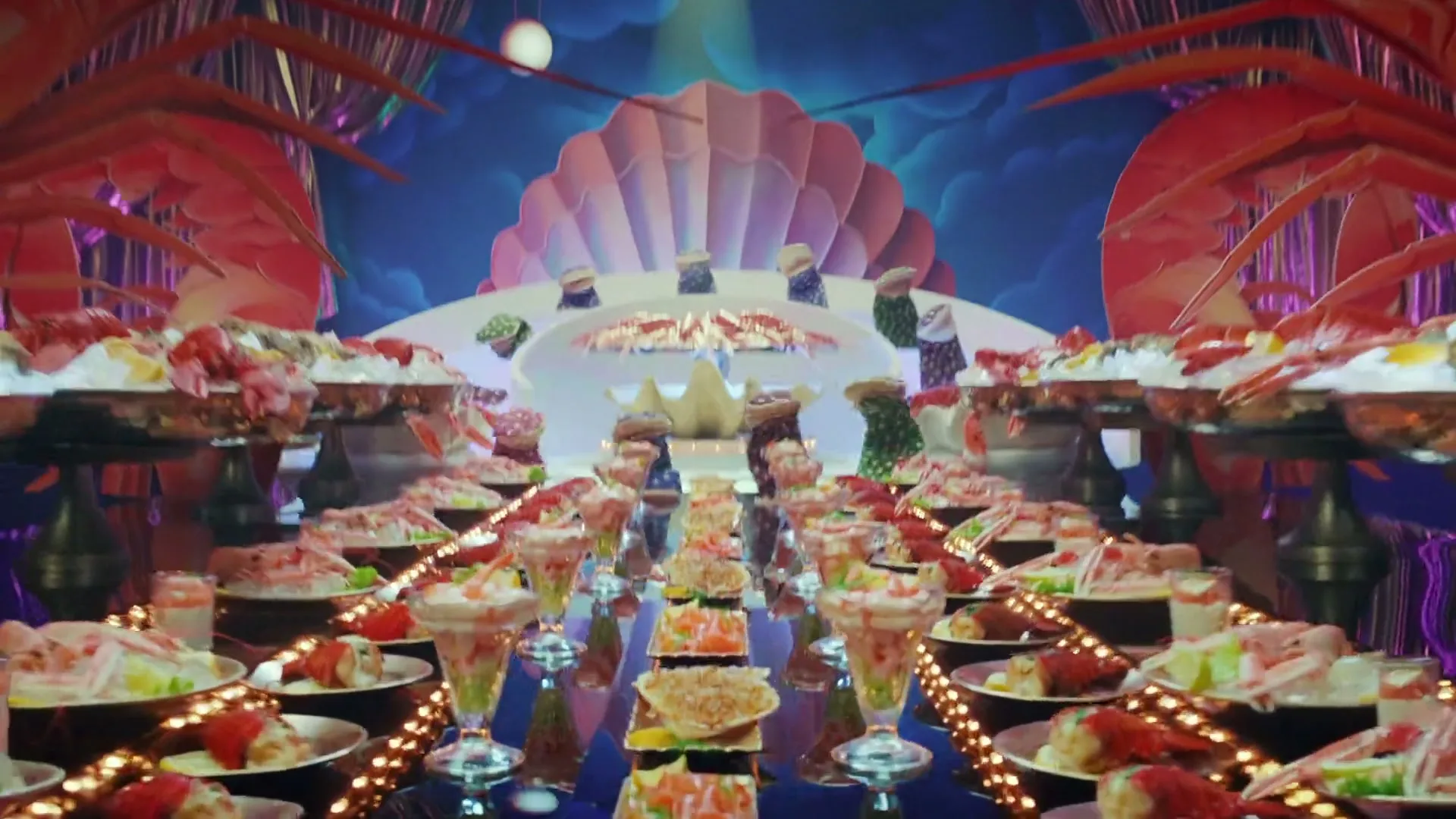 A long table laden with seafood dishes, including prawns, oysters, and crab, is set in a theatrical, purple-draped room with giant prawn cutouts and a seashell backdrop.