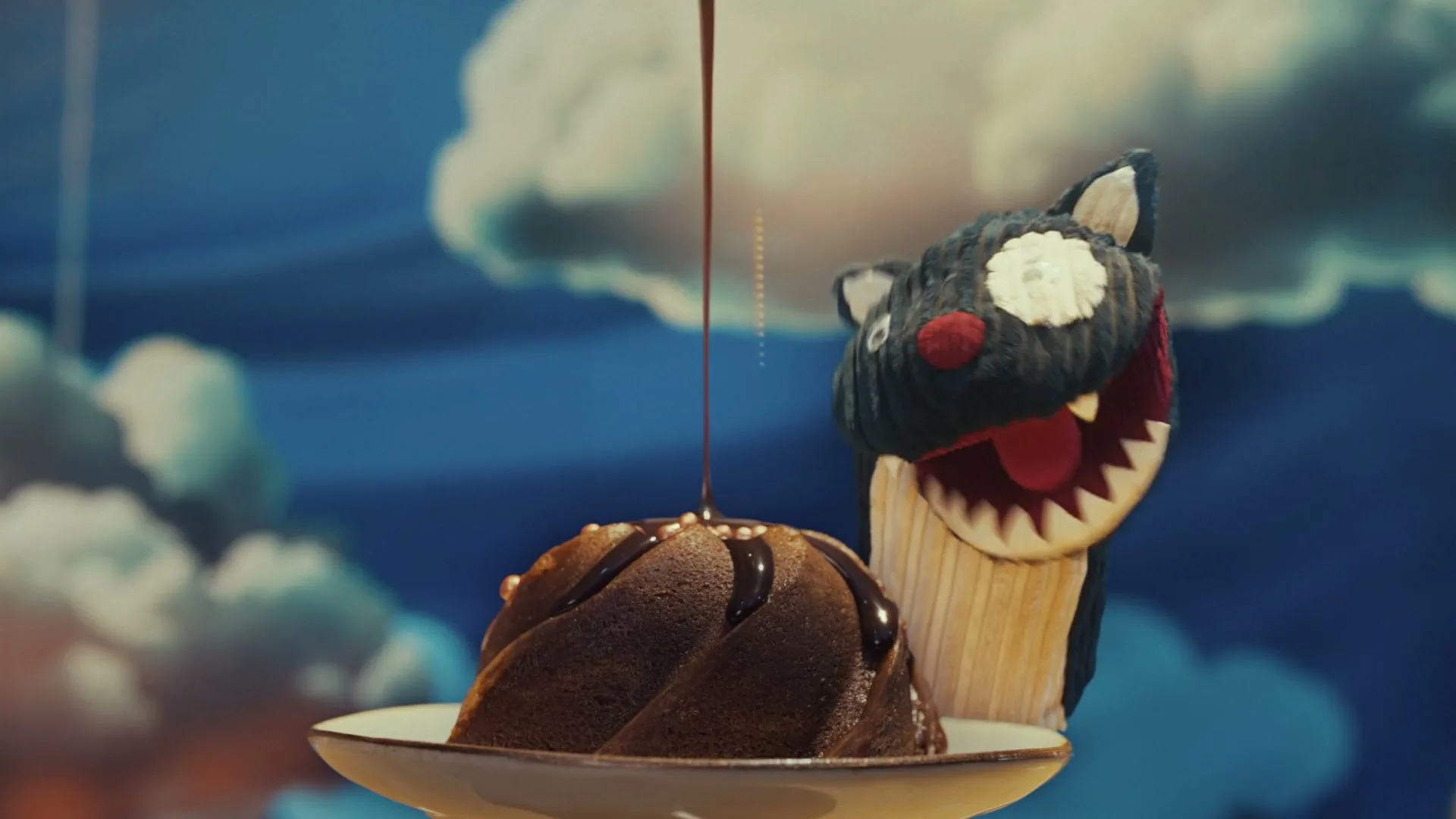 Chocolate sauce is poured onto a bundt cake on a plate, as three colorful monster puppets emerge from behind it against a blue sky backdrop.