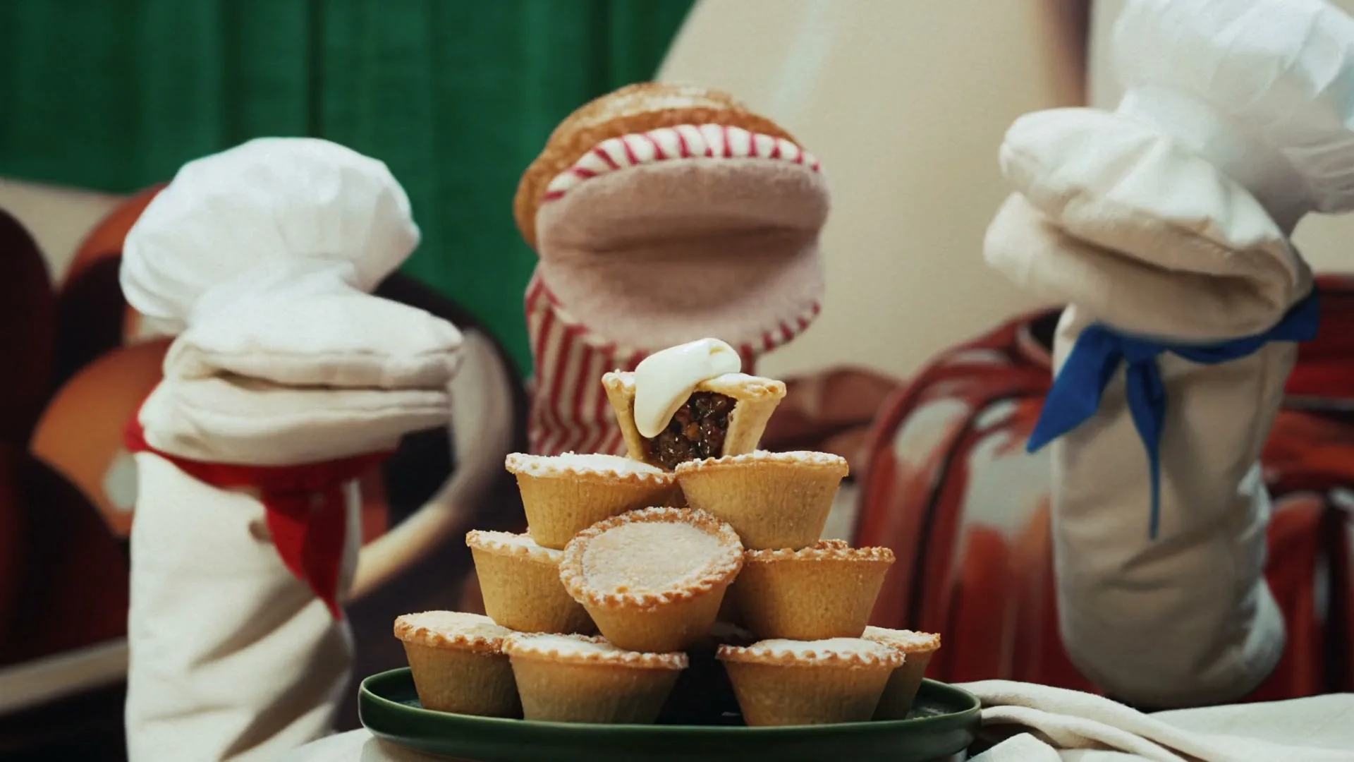 A pyramid of mince pies with one topped with cream is centered, while three oven mitt puppets sing and dance around it in a kitchen setting.