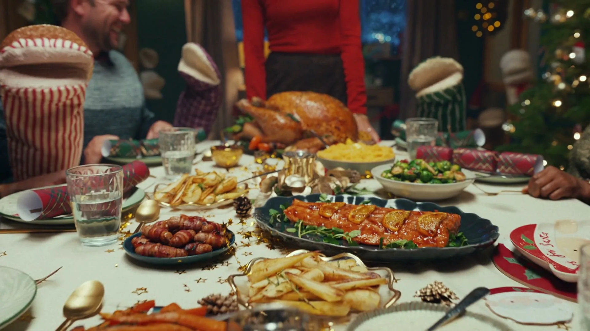A family gathers around a festive Christmas dinner table laden with a roasted turkey, salmon, and various side dishes, with a woman in red standing at the head of the table.