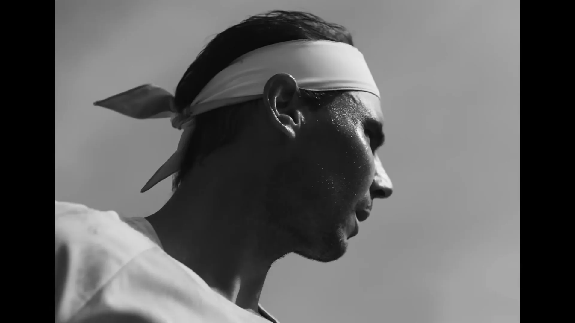 A male tennis player, sweaty and focused, wears a headband and white shirt, looking off-screen in a black and white shot.