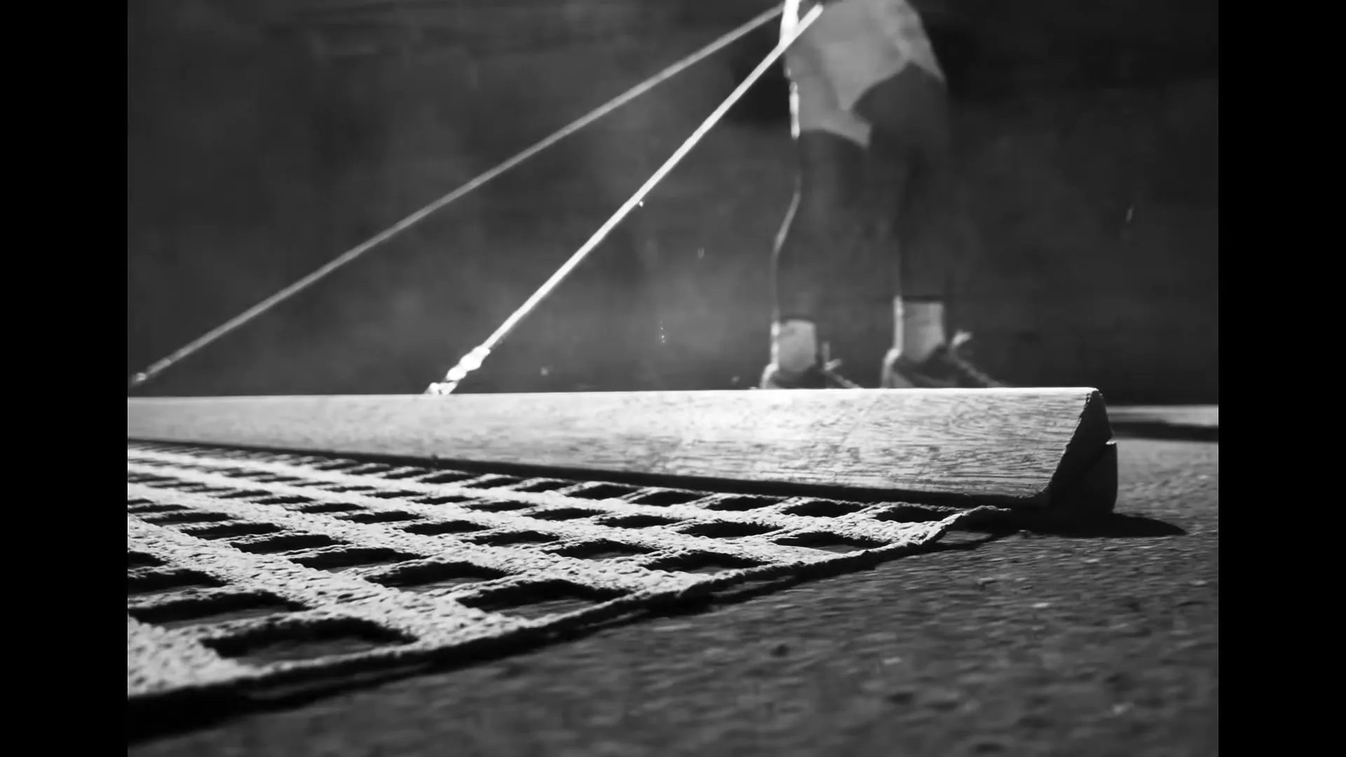 A low-angle, black and white shot shows a person's legs and feet as they walk away from a wooden beam and a textured mat on the ground.