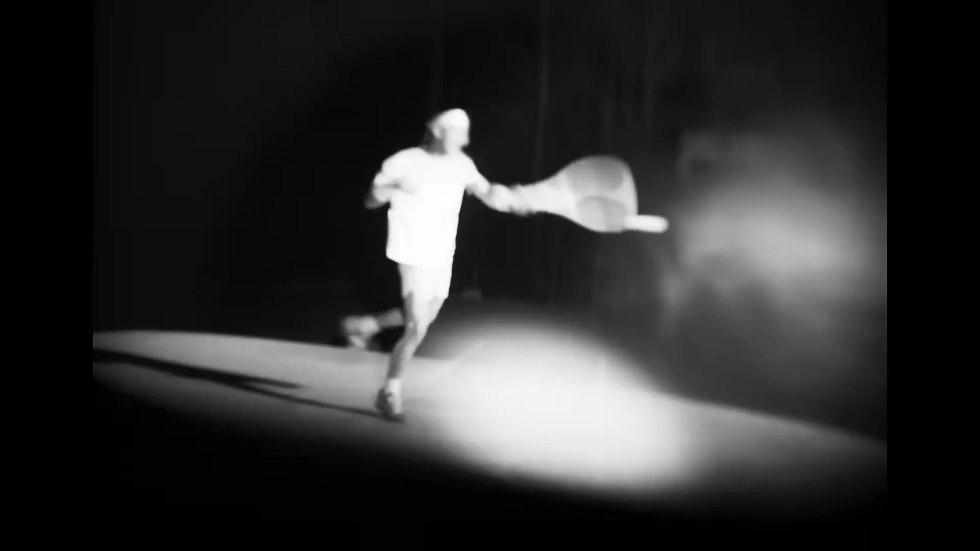 A male tennis player in white sportswear runs across a dark tennis court, swinging his racket in a low-key, black and white shot.