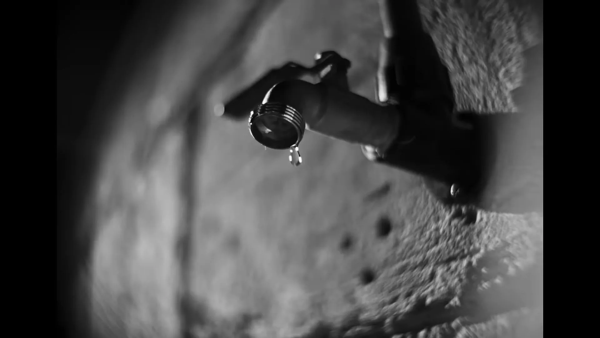 A black and white extreme closeup shot of a dripping faucet on a textured wall, emphasizing the slow fall of water.