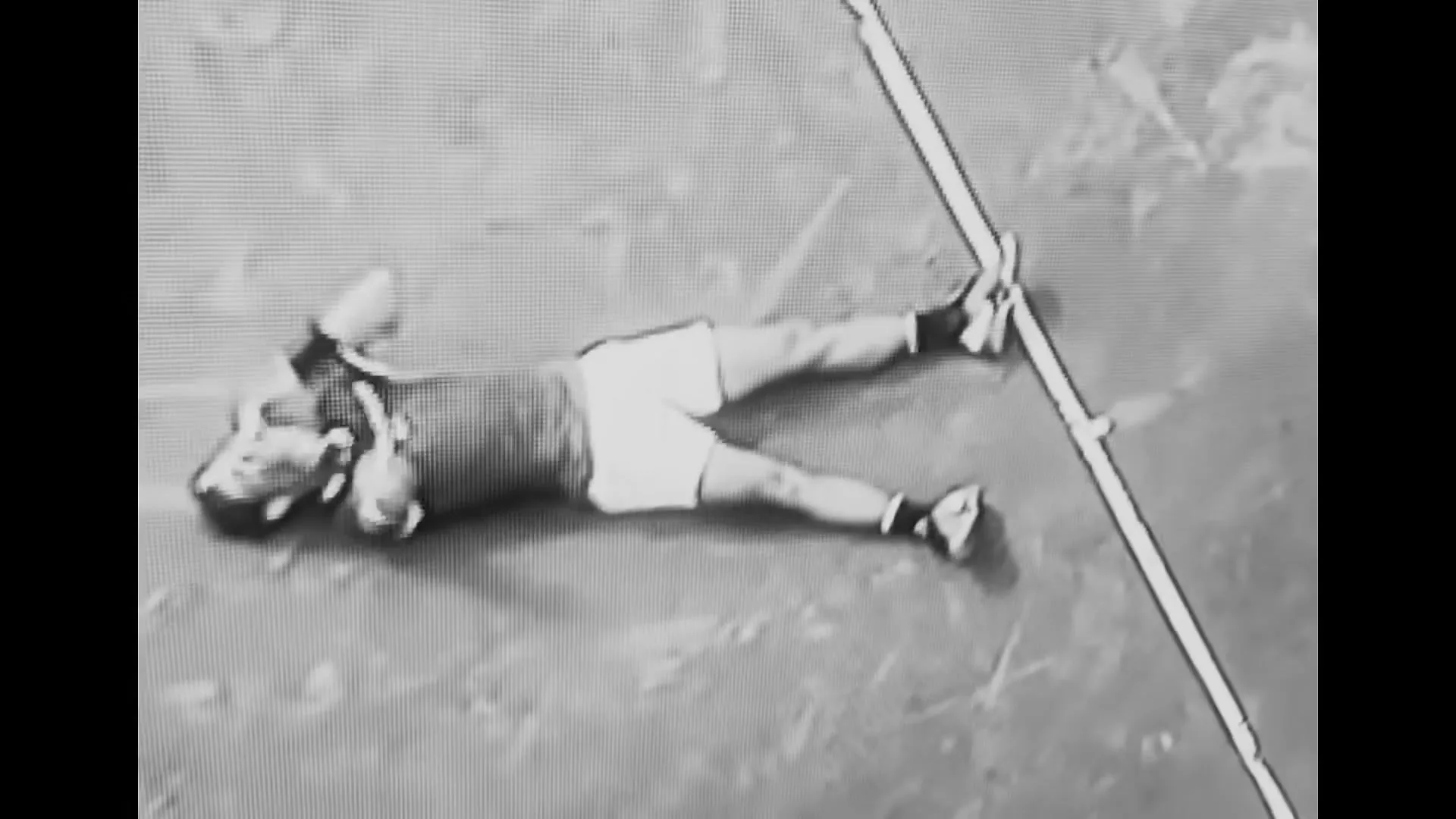 A male athlete in a tank top and shorts lies on a court, stretching his arms and legs, in a black and white archival shot.