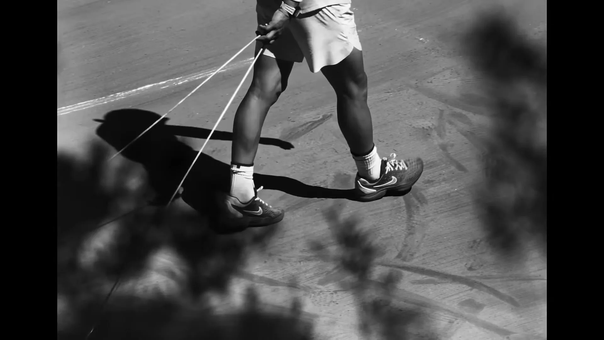 A person in athletic shorts and sneakers jumps rope on a concrete surface, captured in black and white with strong shadows.