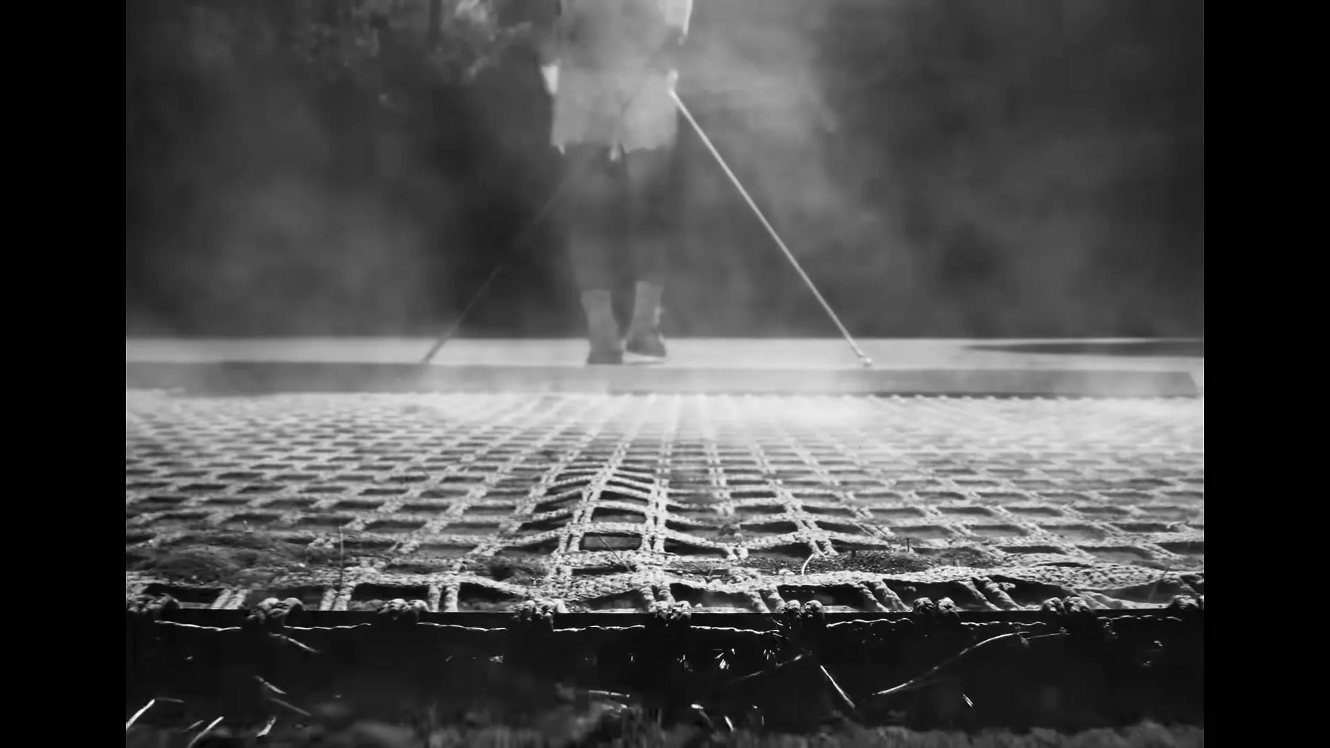 A black and white low-angle shot shows a person's legs walking on a textured surface, with smoke rising around them in an outdoor setting.