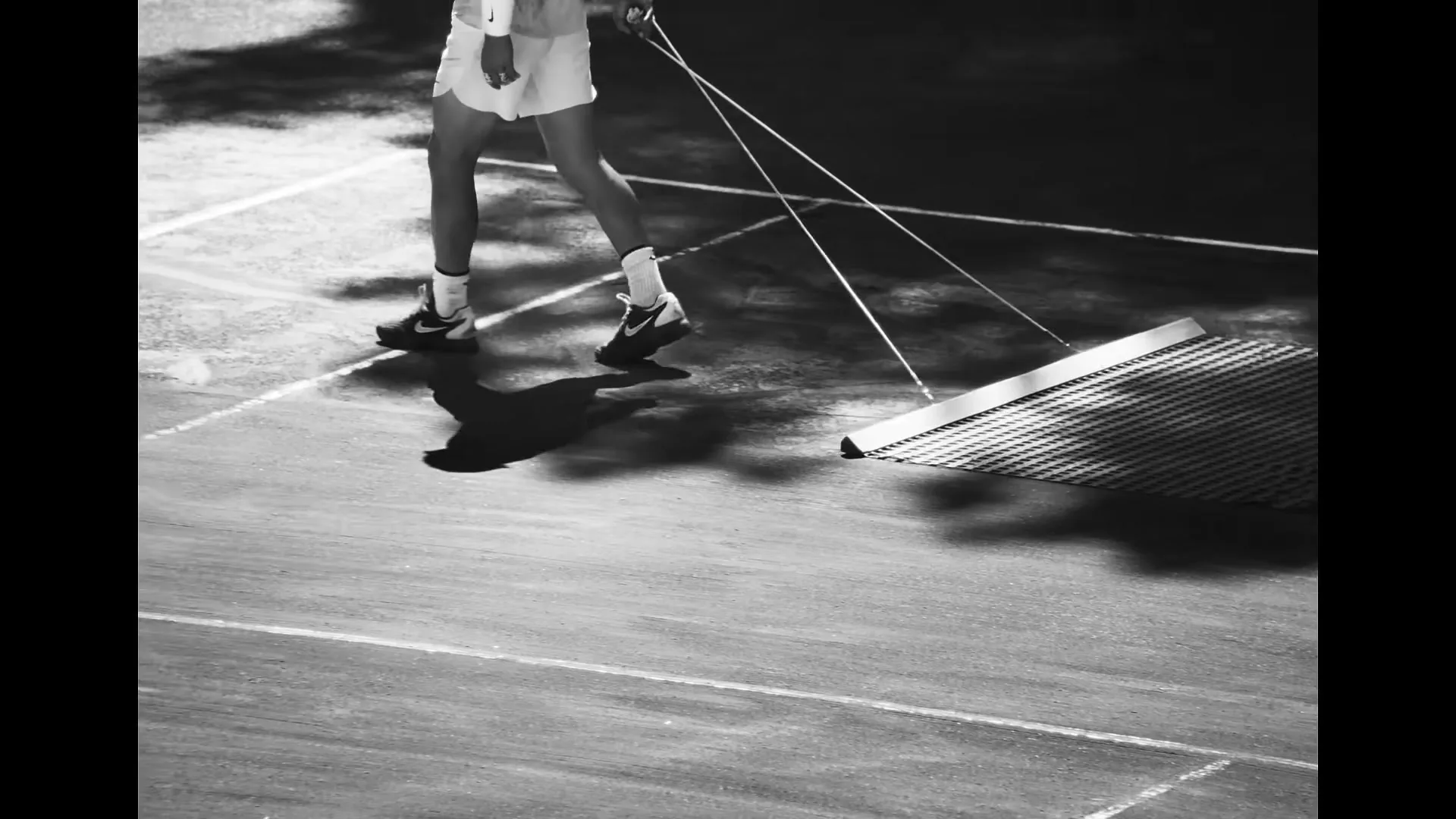 A tennis player in shorts and sneakers drags a court sweeper across a sun-dappled tennis court, shot in black and white.