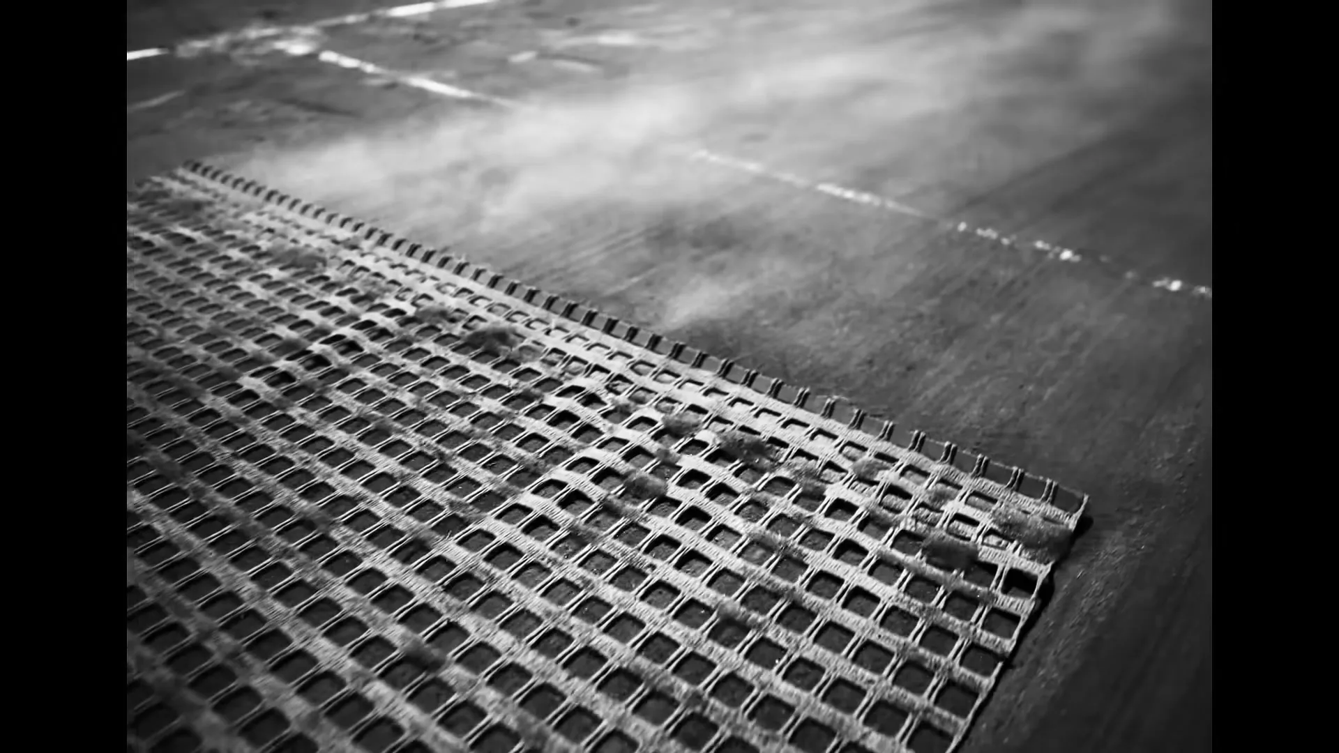 A close-up, low-angle shot shows a metal grate on a wet, dark surface with steam rising, in black and white.