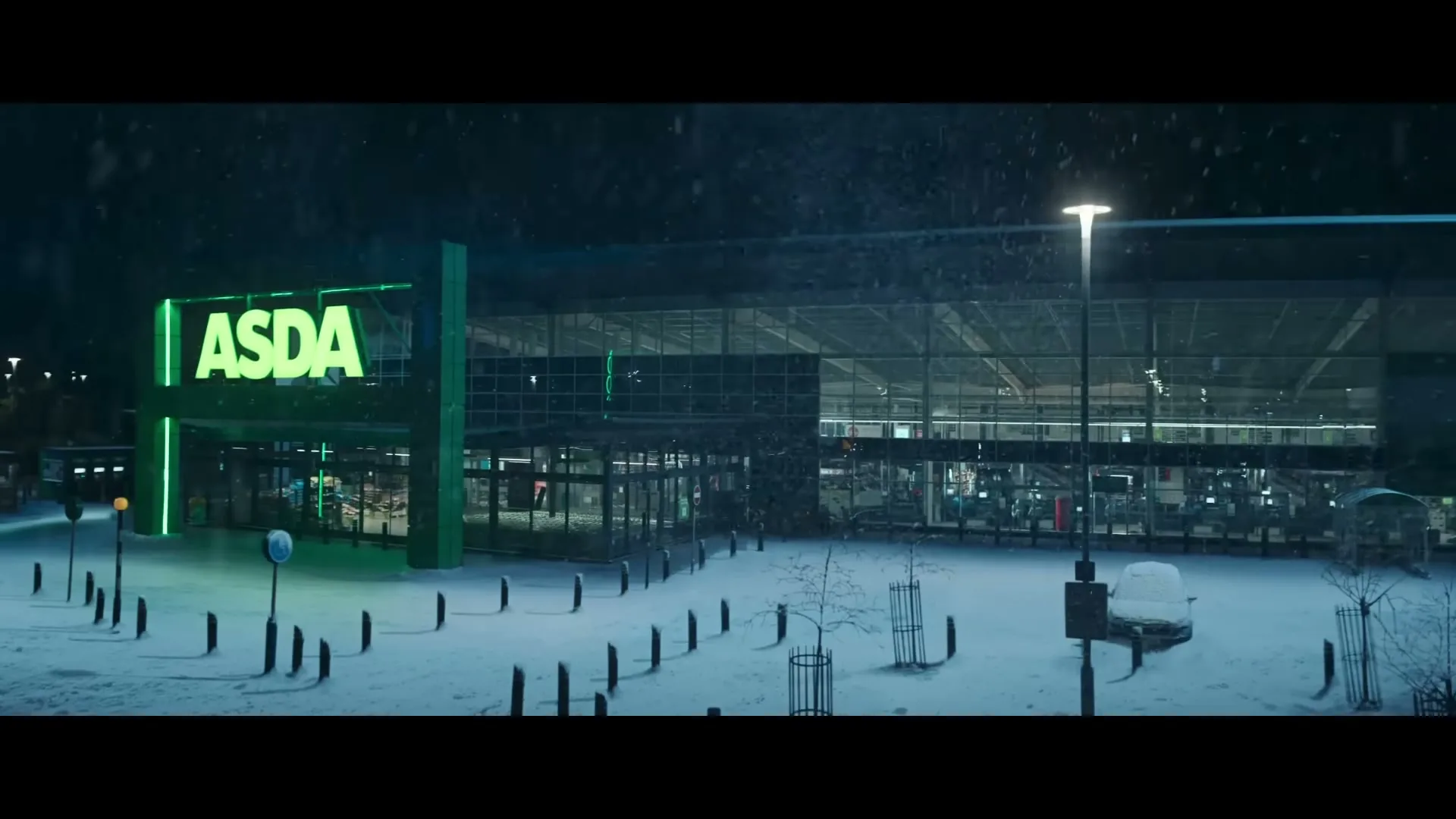 An exterior wide shot of an Asda supermarket at night, with snow falling and covering the ground.