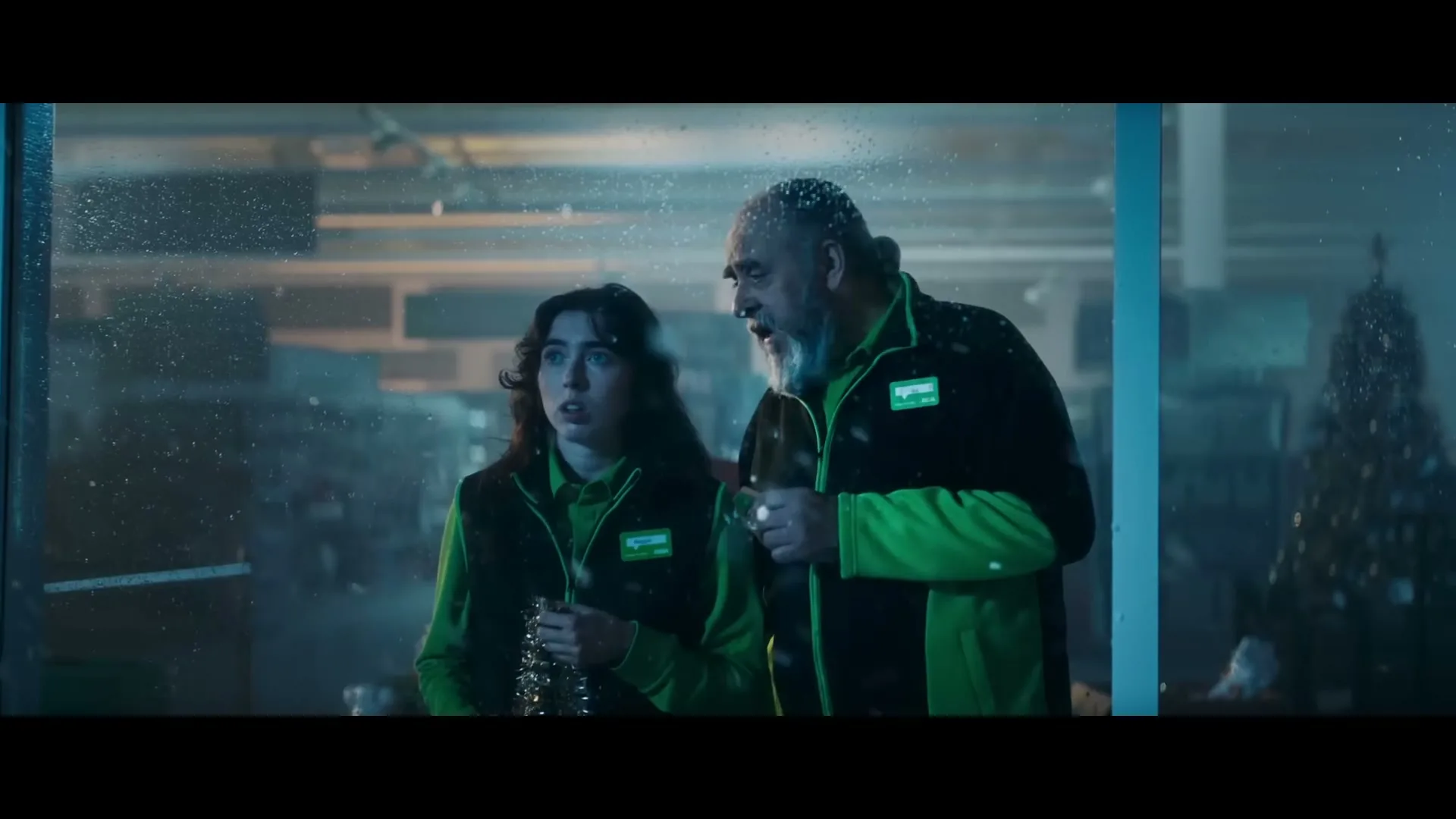 A young female and an older male Asda employee look out a snowy window at night, both wearing green and black uniforms.