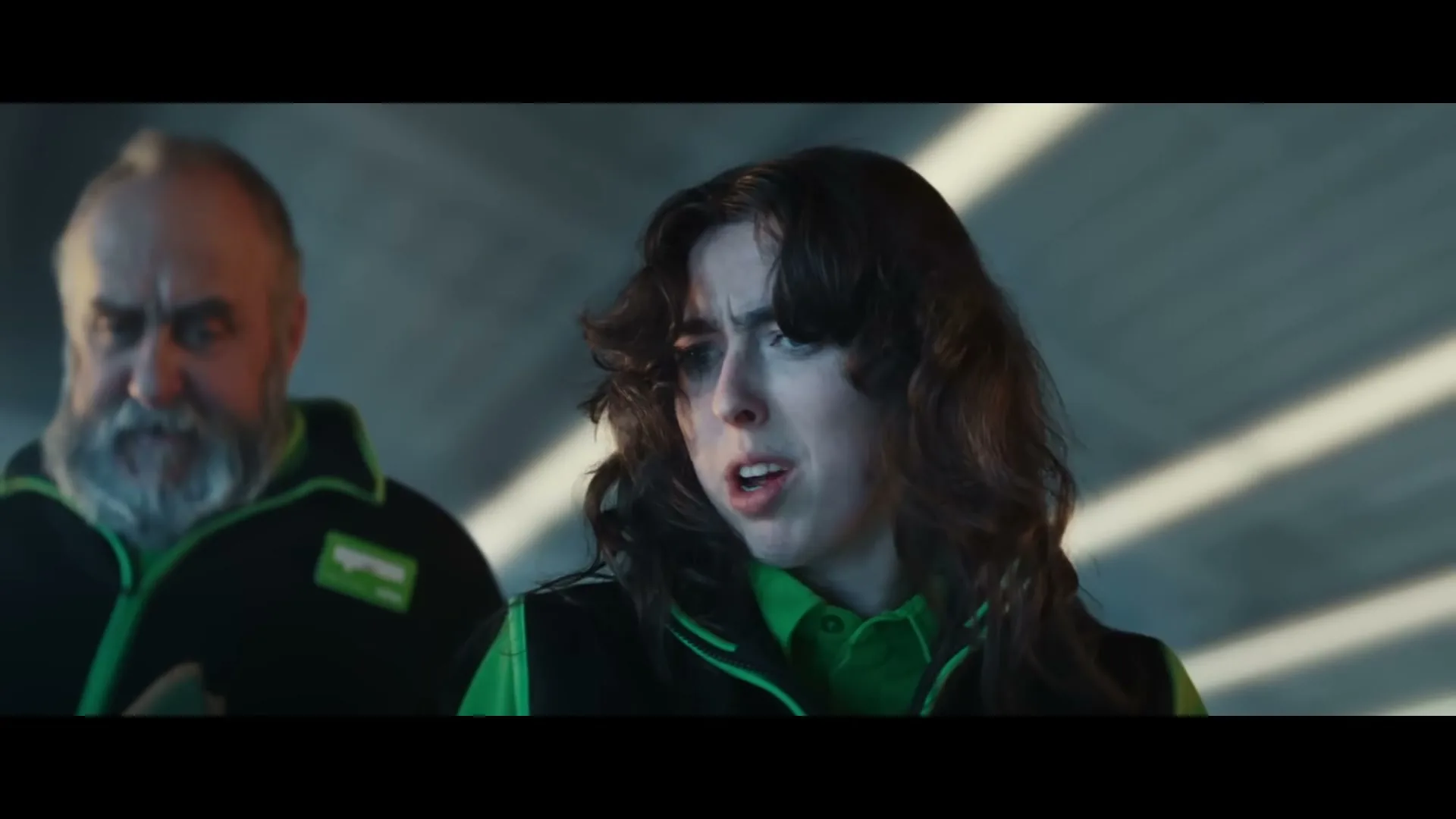 A young woman with dark hair and a man with a white beard, both wearing green and black uniforms, look surprised in a parking garage.