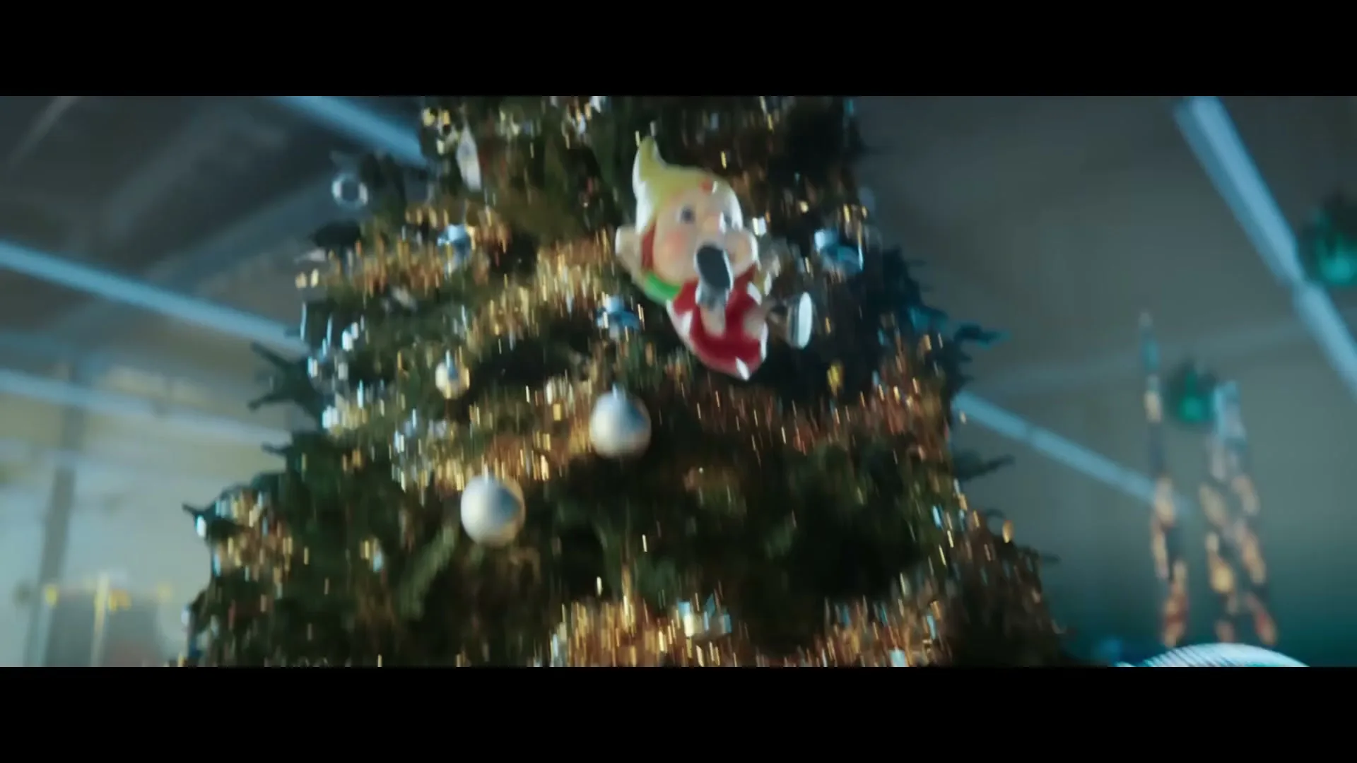 A small animated elf with a red and white outfit and a yellow hat is seen falling down a large, decorated Christmas tree in a warehouse.