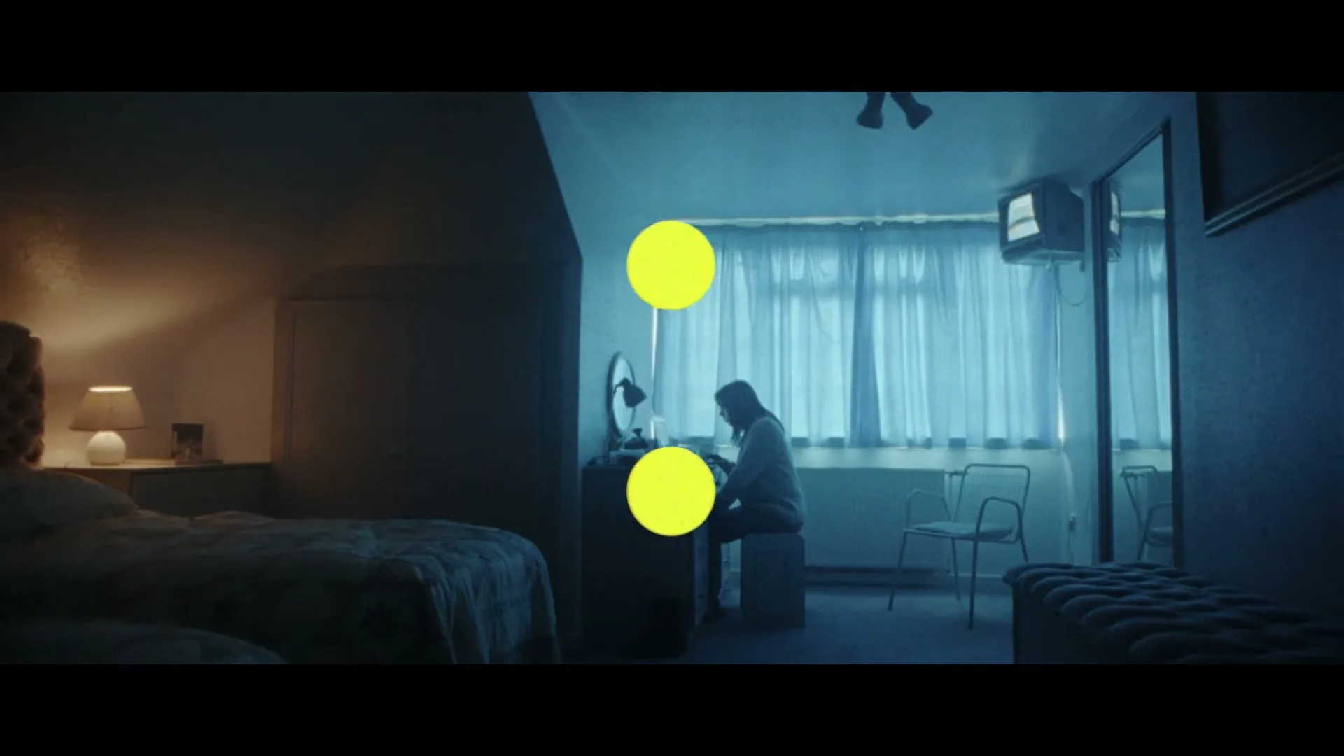 A young woman sits at a dressing table in a dimly lit bedroom, illuminated by a blue light from the window, as yellow dots appear on screen.
