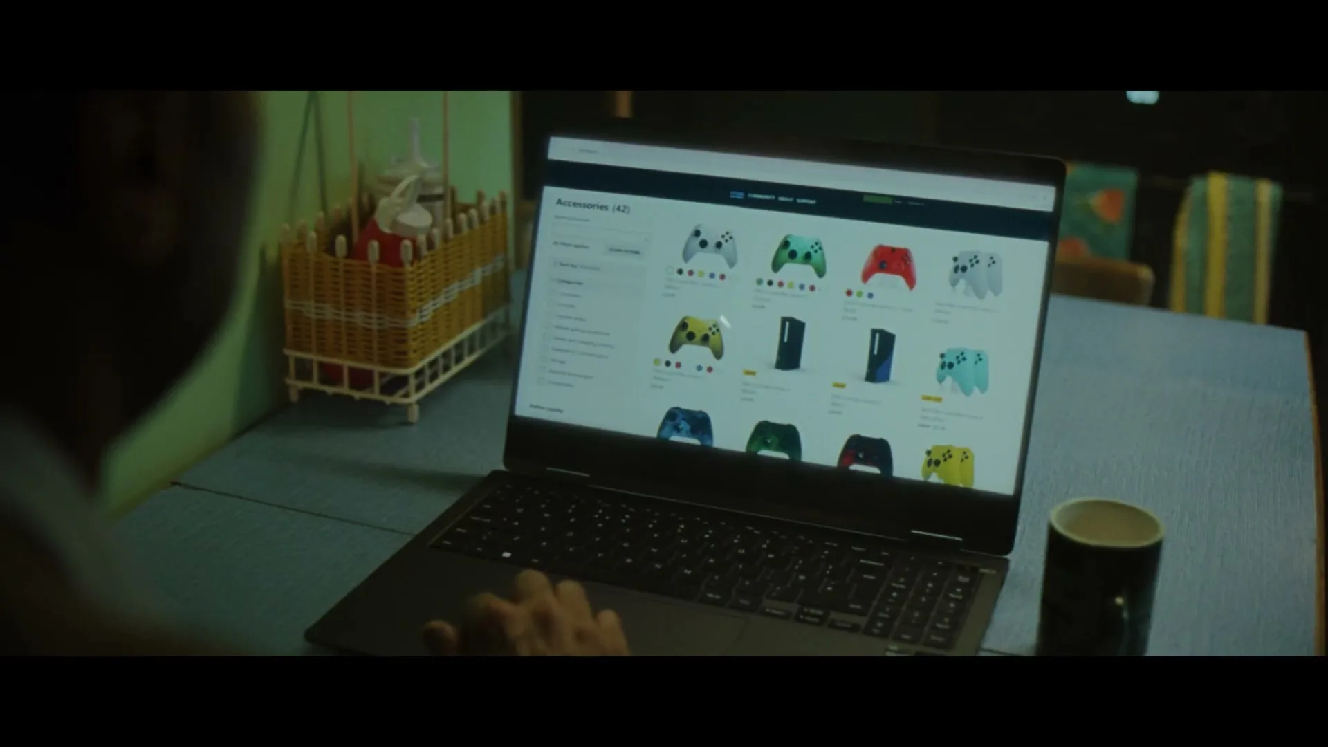 A person's hands are seen typing on a laptop, which displays a website selling gaming accessories, on a table in an interior setting.