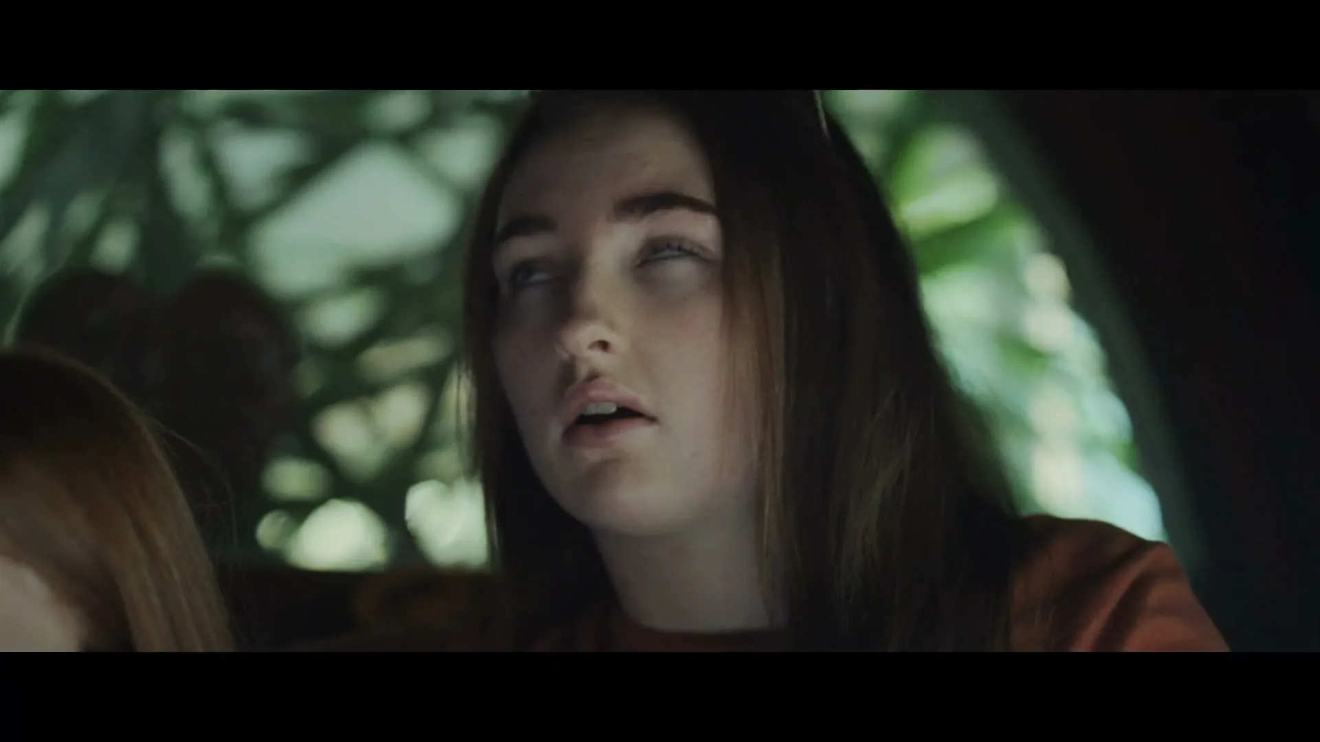 A young woman with long brown hair and freckles looks up with wide eyes, then closes them, in a dark, green-lit interior.