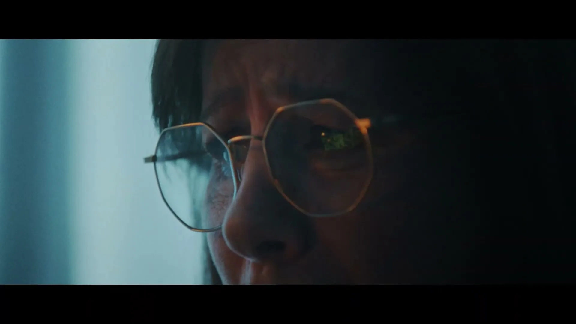 A person with dark hair and glasses looks intently, with a reflection of green text visible in their glasses, in a dimly lit interior.
