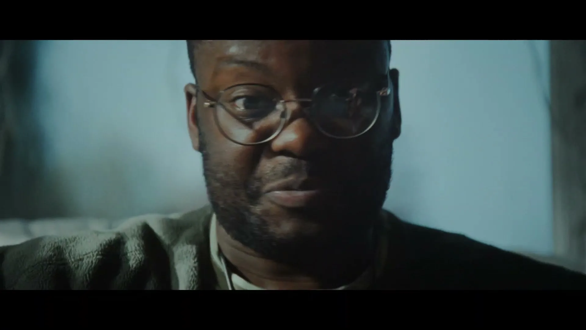 A Black man with glasses looks directly at the camera, speaking with a concerned expression in a dimly lit interior.