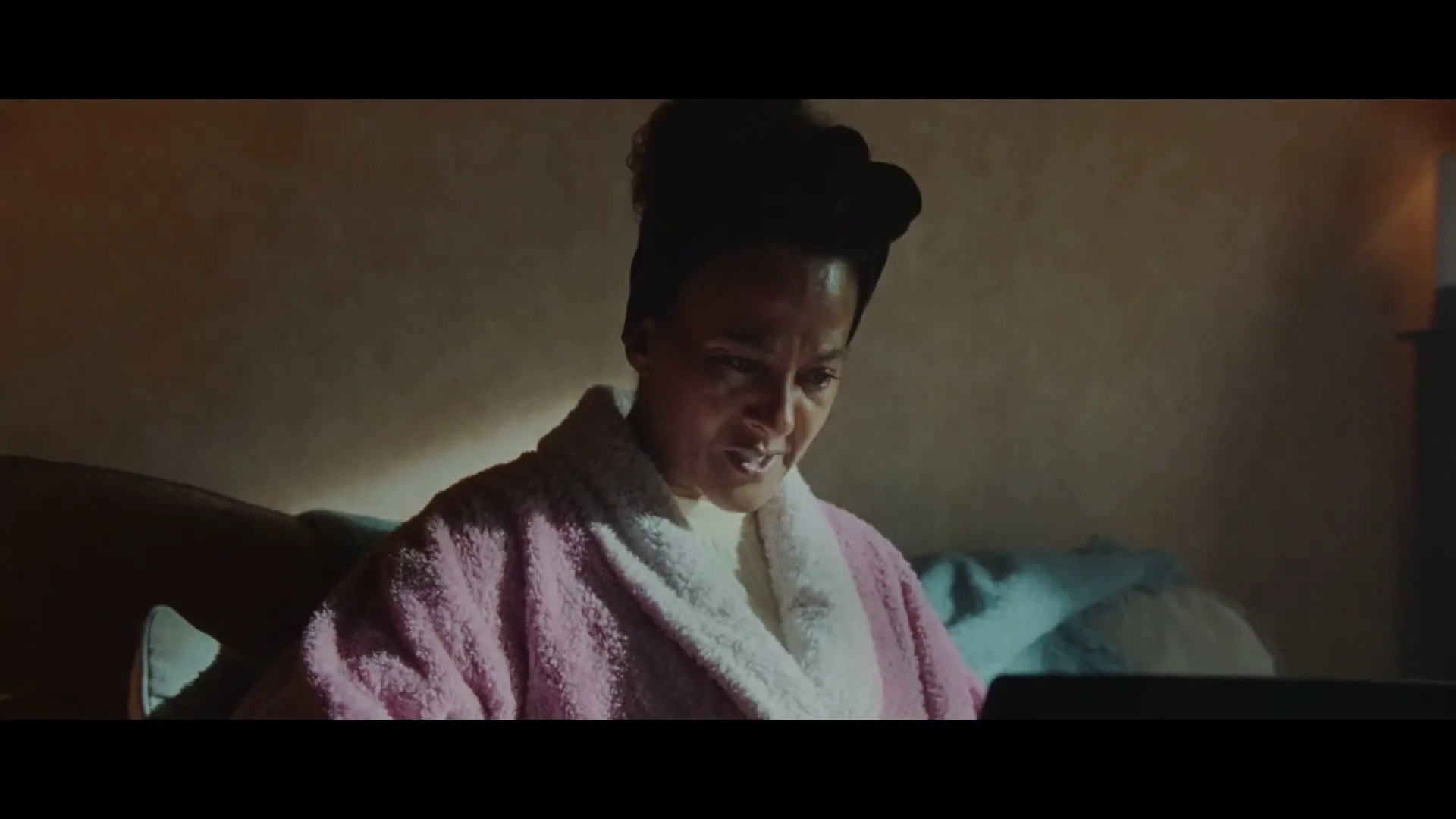 A woman in a pink robe looks distressed while using a laptop, then the camera pans to a framed photo of a smiling boy.