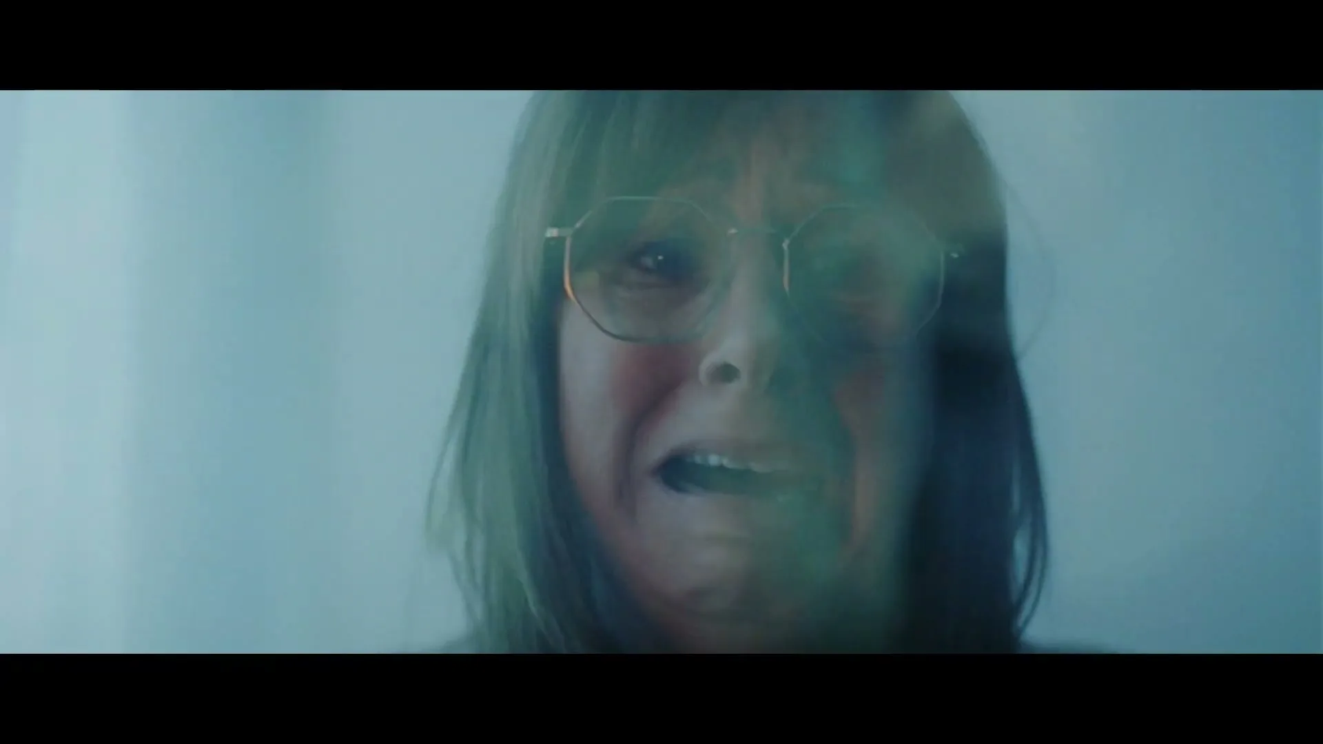 A woman with glasses and brown hair is seen in a medium closeup, crying and distressed, with reflections on a glass surface in front of her.