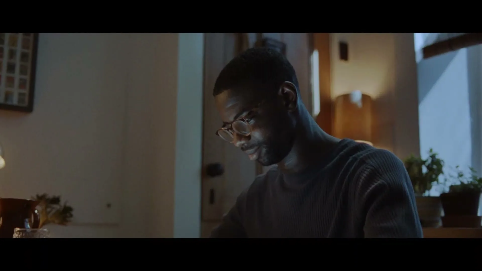 A man with glasses looks down, illuminated by a warm light, in a dimly lit room with plants in the background.