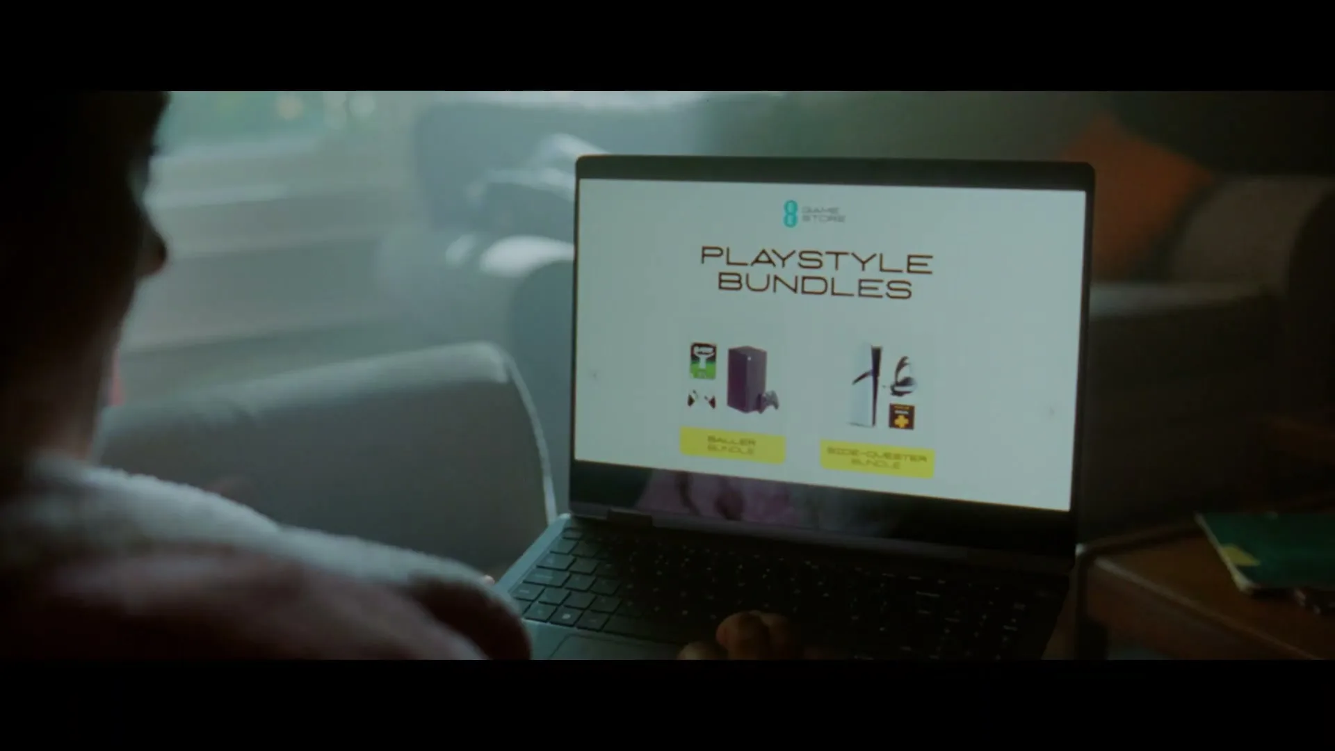 A person is seen from behind, looking at a laptop screen displaying "PLAYSTYLE BUNDLES" with images of gaming consoles, in a dimly lit living room.