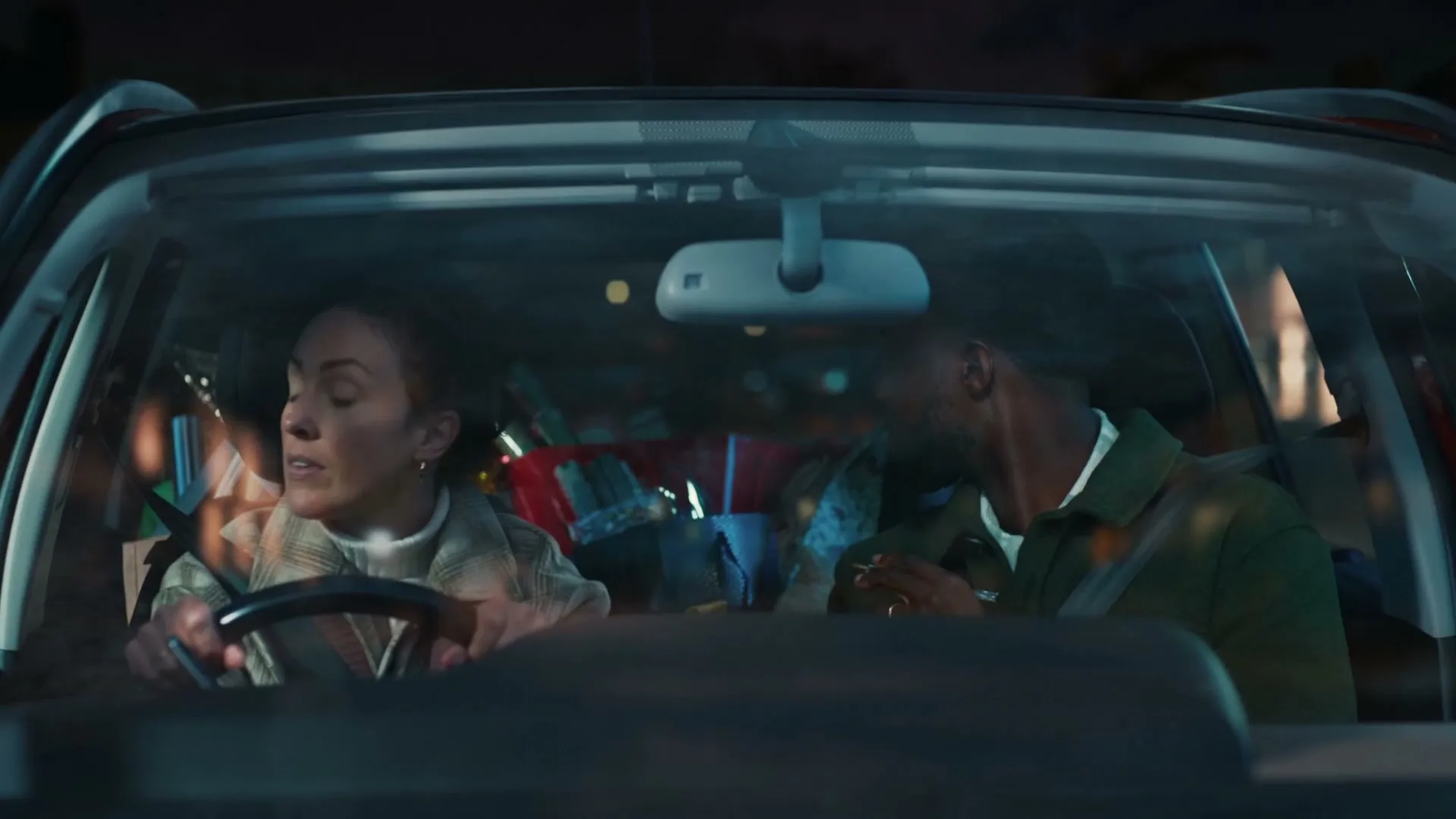 A woman drives a car at night, looking concerned, while a man in the passenger seat looks down, with Christmas gifts visible in the back.