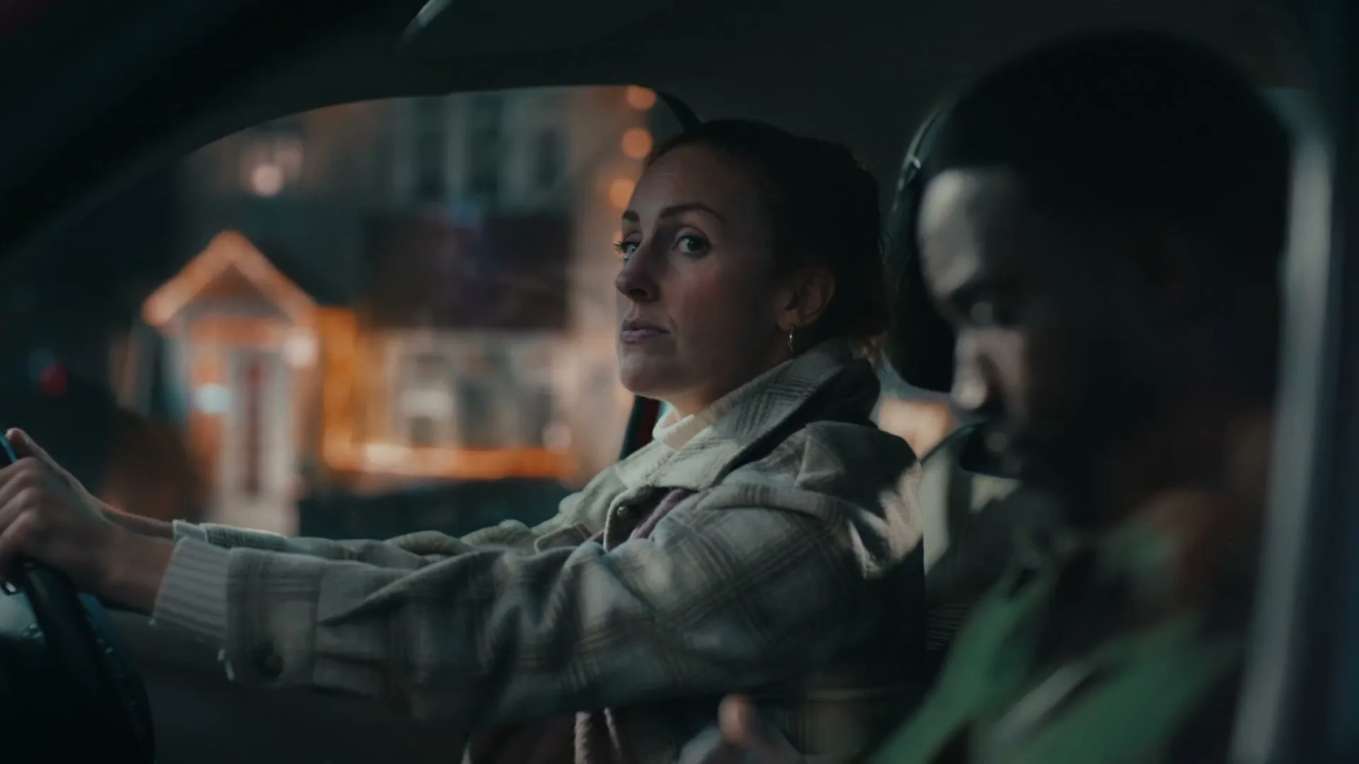 A young woman with red hair drives a car at night, looking at the road, while a man sits in the passenger seat.