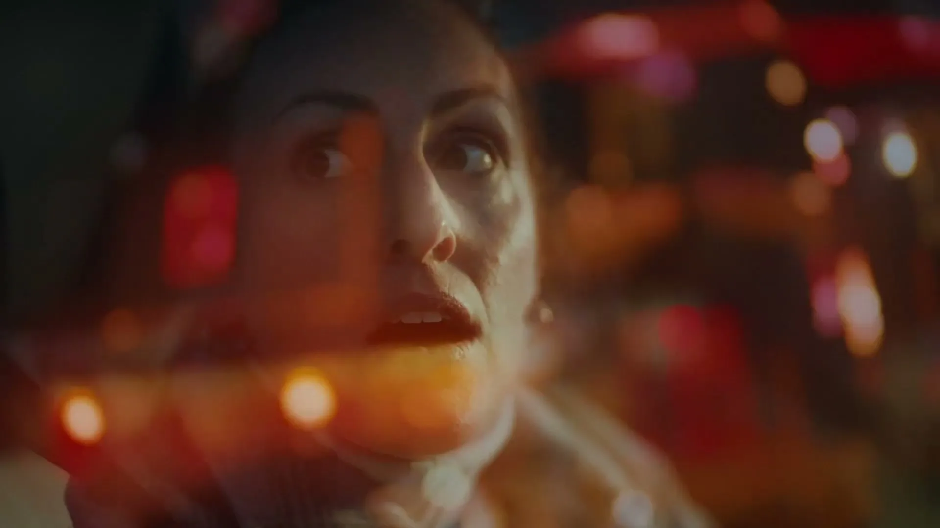 A woman with dark hair and a white turtleneck looks surprised, with blurred red and orange lights reflected on her face, in a car at night.