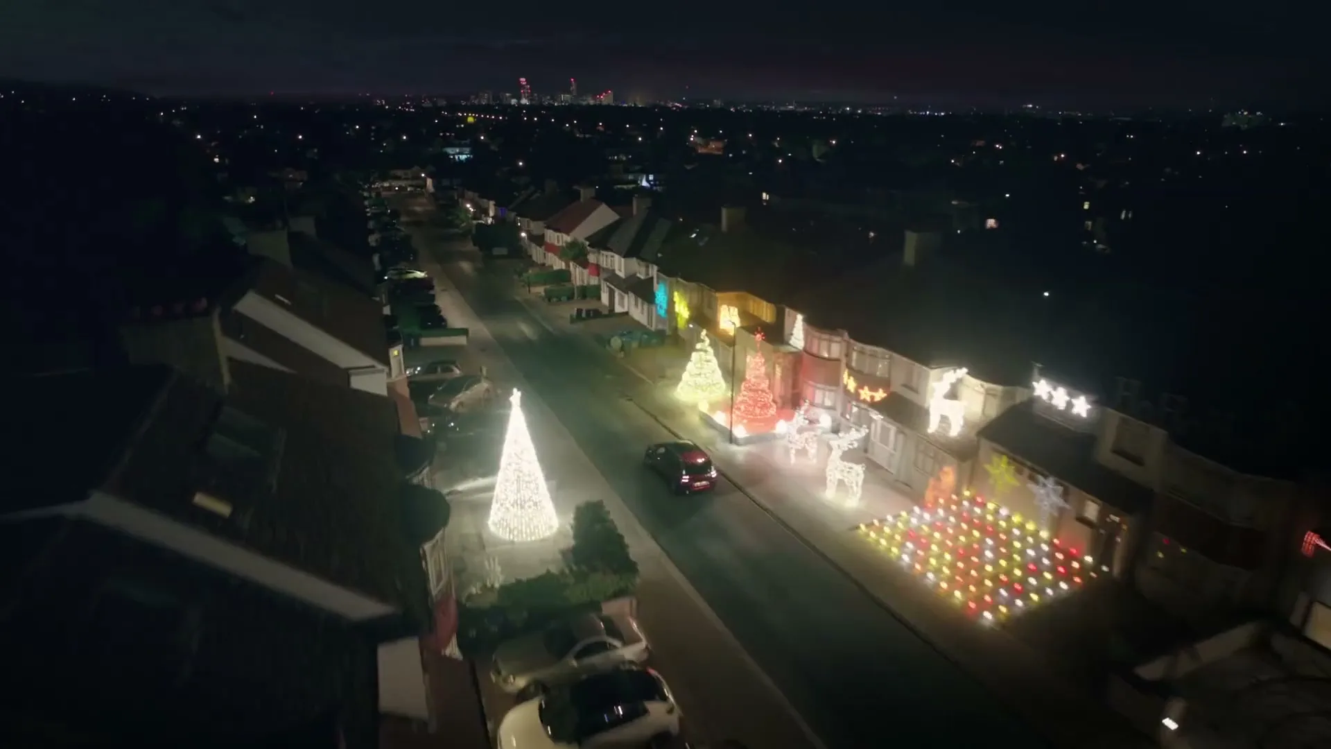 An aerial view of a suburban street at night, with houses decorated with elaborate Christmas lights, including a large "HoHoHo" sign.