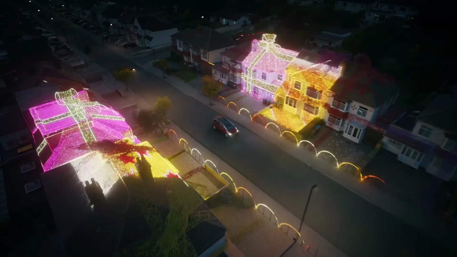 An aerial shot shows a car driving down a street at night, with houses on both sides decorated with colorful Christmas lights that resemble wrapped gifts.