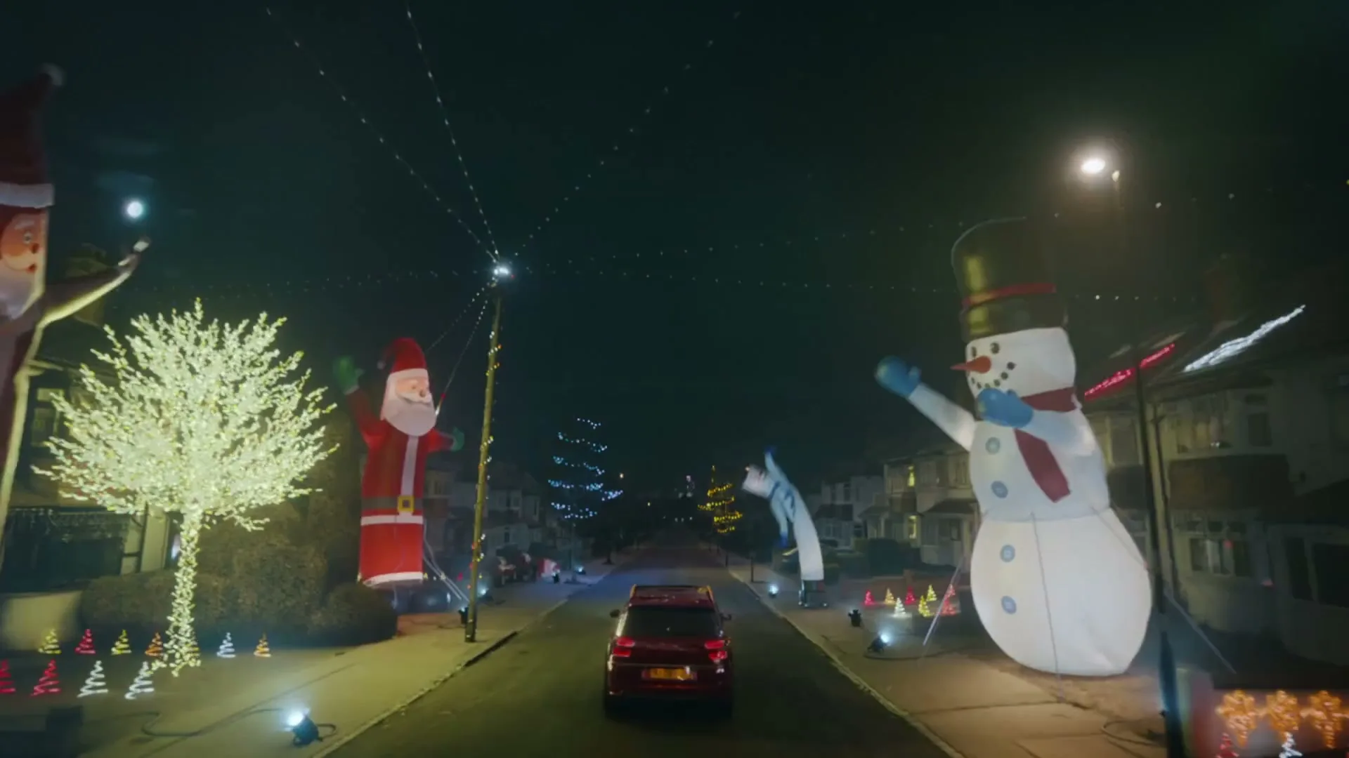 A red car drives down a suburban street at night, surrounded by houses decorated with elaborate Christmas lights and large inflatable Santa Clauses and snowmen.