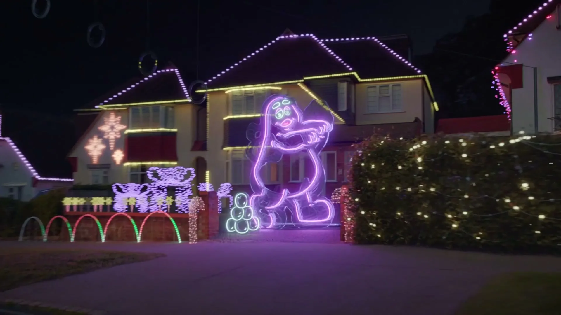 A suburban house is decorated with elaborate Christmas lights, including a large, glowing purple Grimace figure, at night.