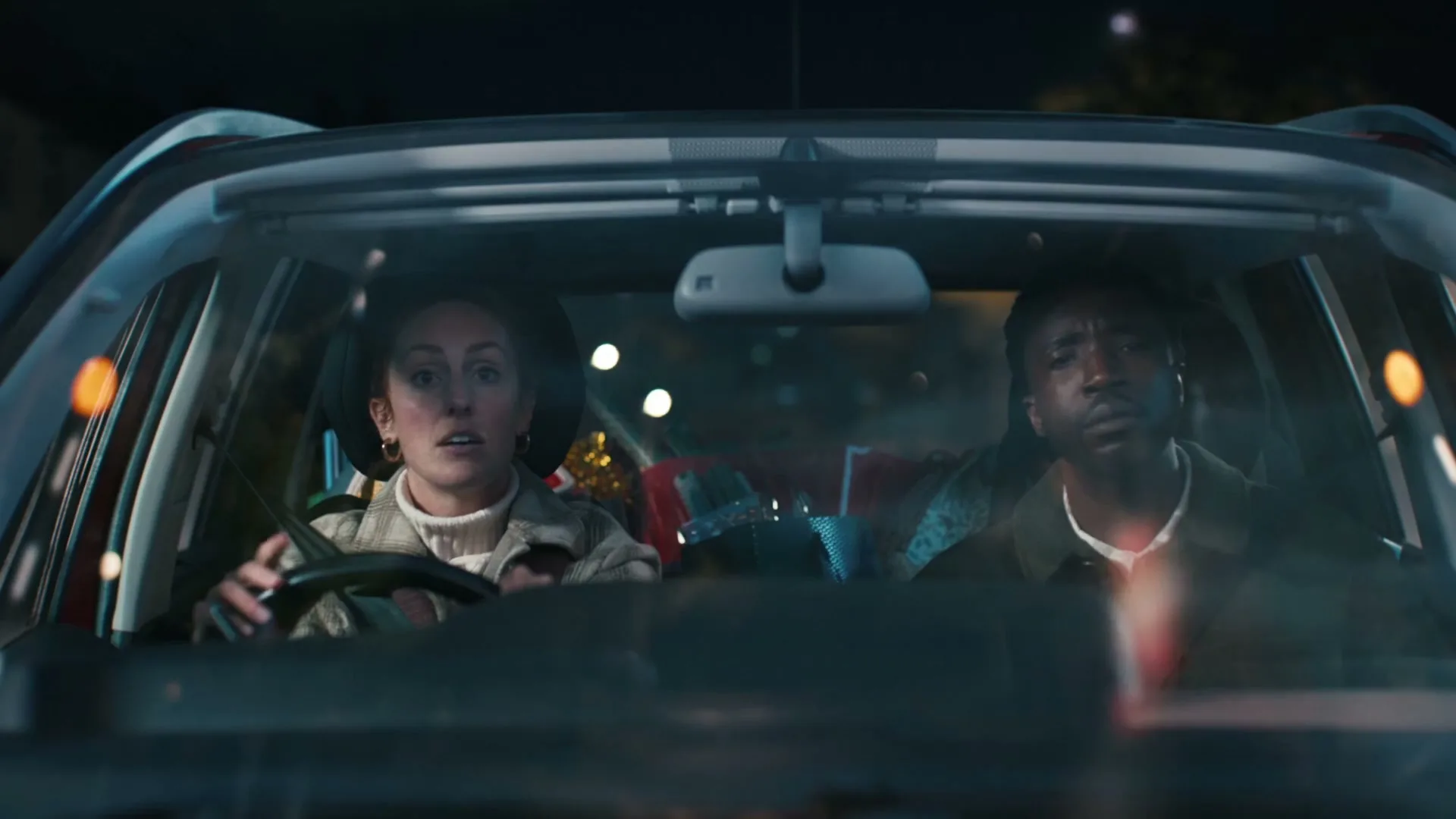 A woman and a man are seen through the windshield of a car, driving at night with Christmas presents in the back seat.