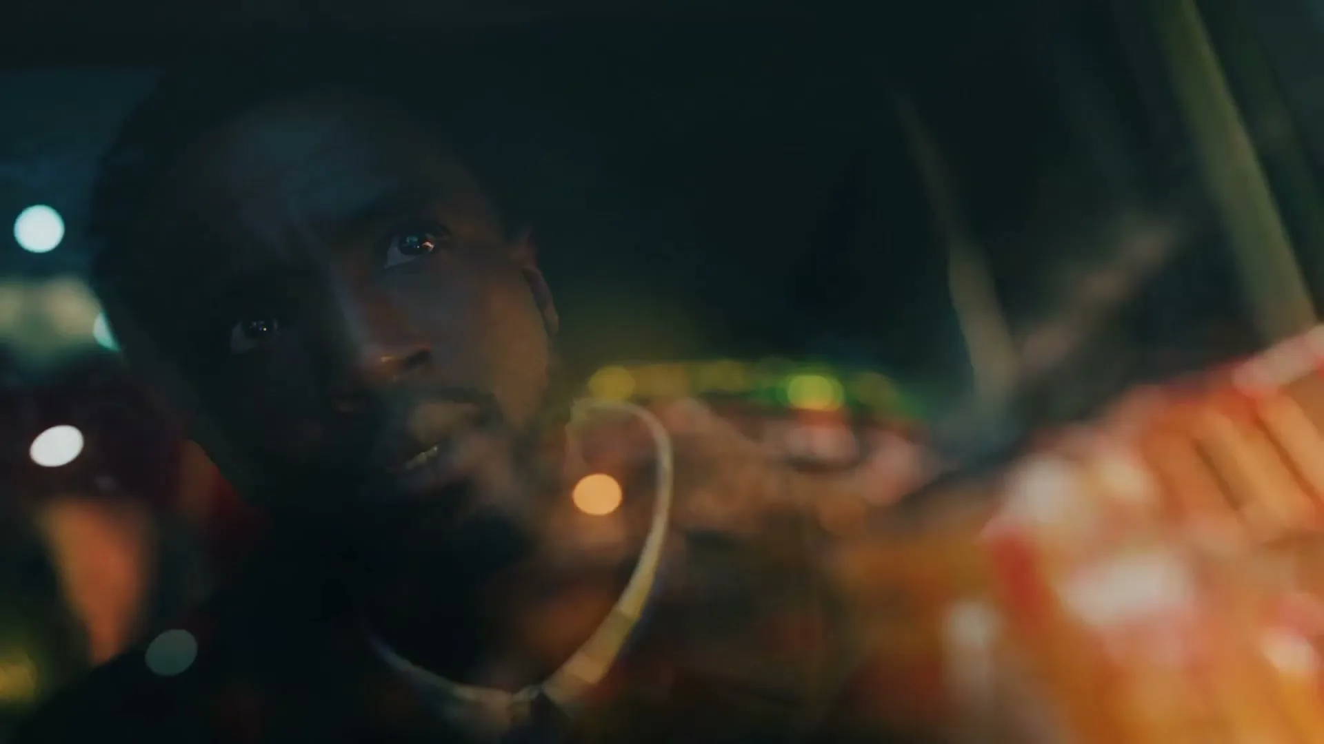 A man with a beard looks up and to the right, with blurred lights reflecting on a window in the foreground at night.