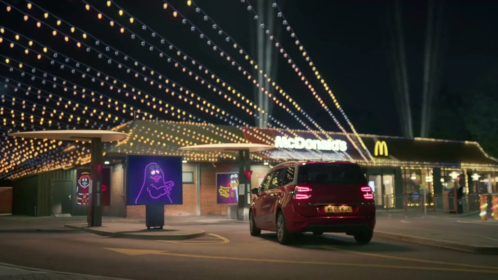 A red car drives through a McDonald's drive-thru at night, decorated with festive lights and animated characters on digital screens.