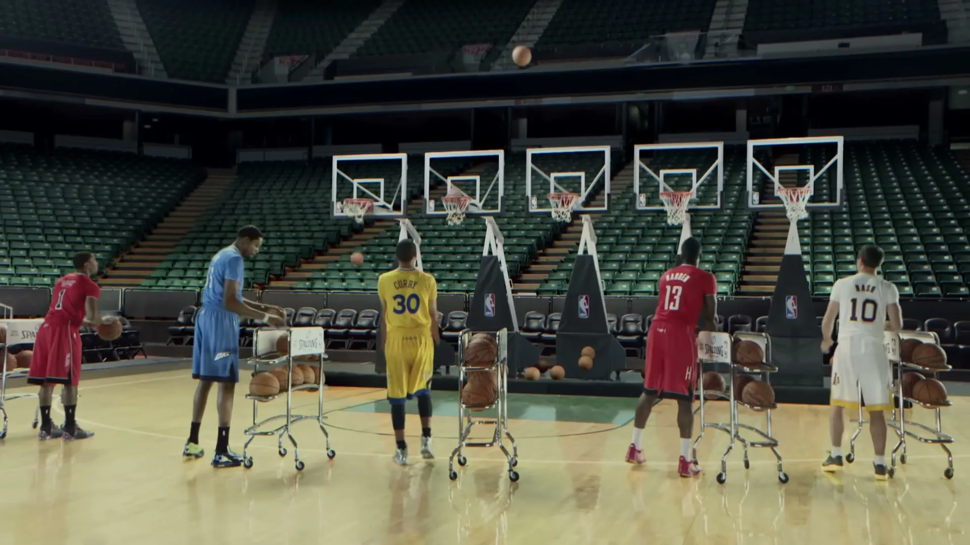 Five NBA players in their team jerseys shoot basketballs at multiple hoops in an empty arena.