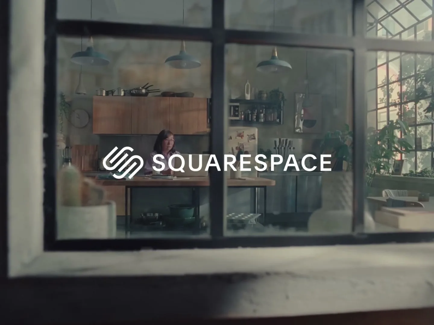 A woman with dark hair and glasses sits at a table in a rustic kitchen, working on a laptop, viewed through a window with the Squarespace logo overlaid.